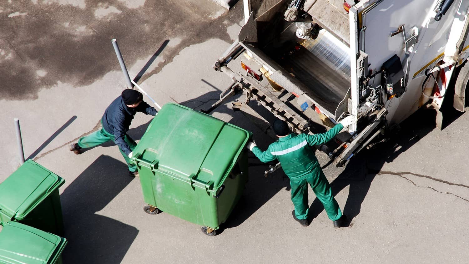 A person wearing protective gear collecting waste in to a truck, with garbage bins in the foreground.