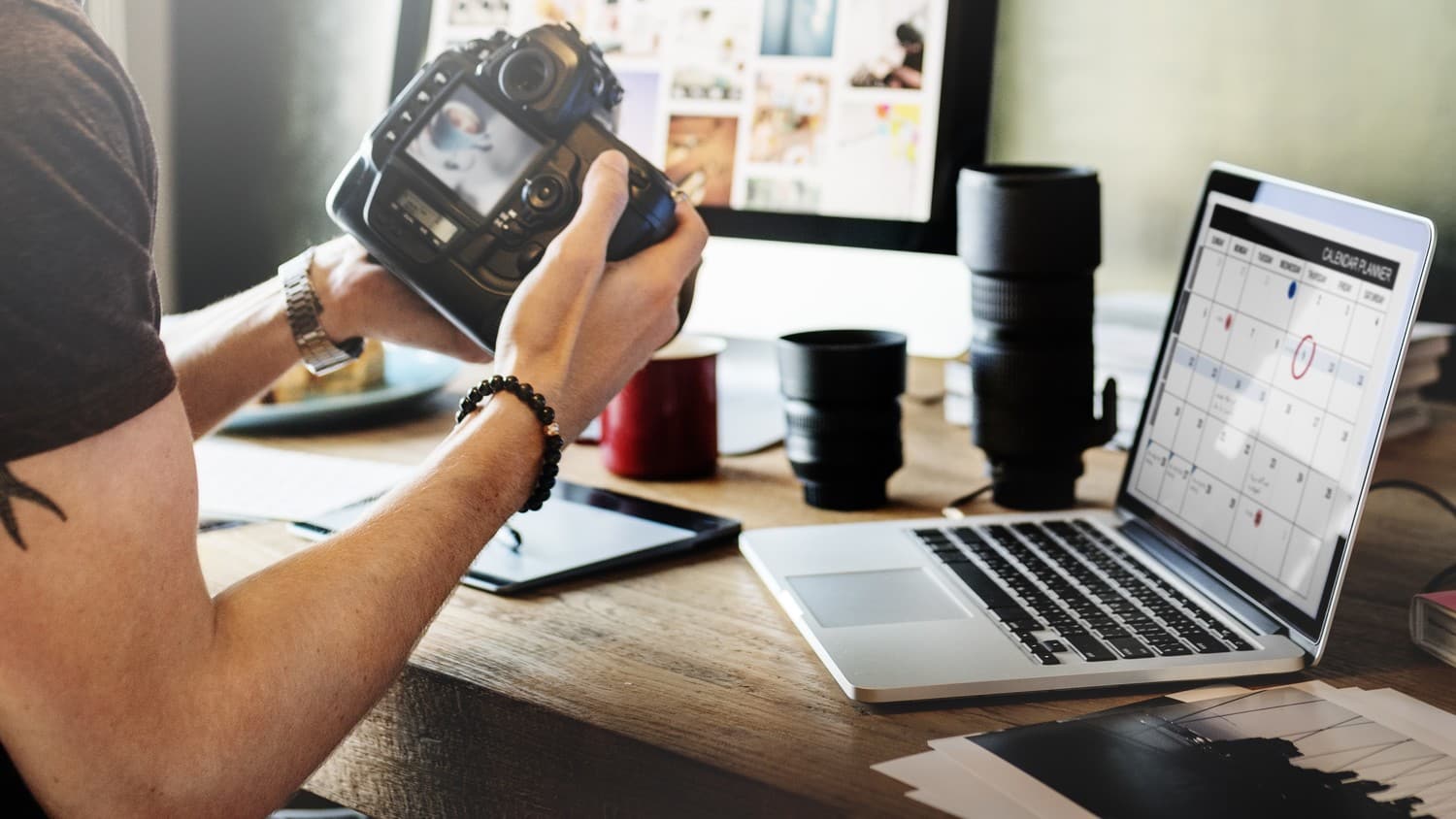 A photographer holding his camera in frontt of his desk, surrounded by his computer, lenses, and other equipment.