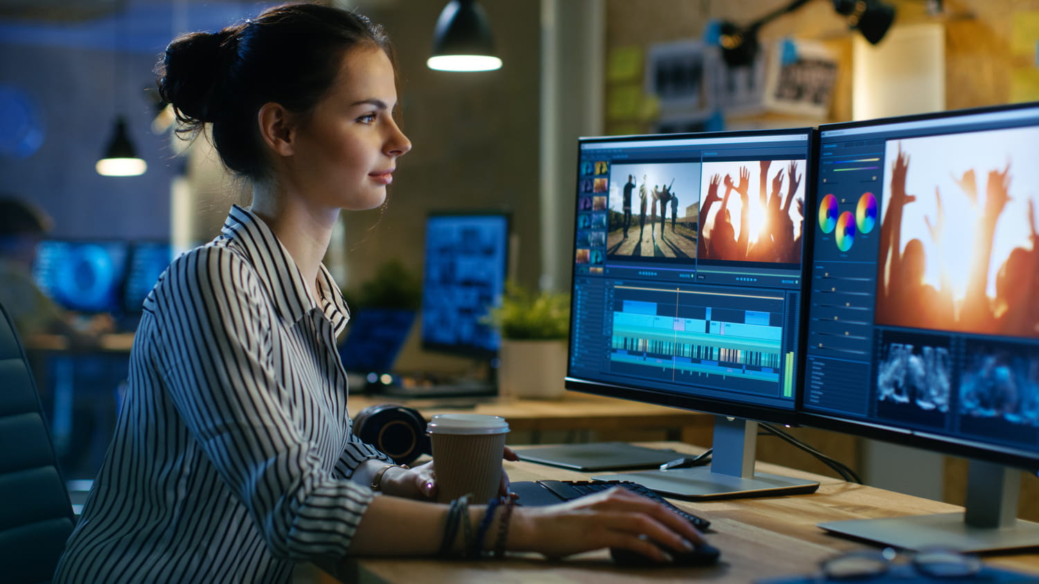 A girl with a coffee seated on her office desk with 2 monitors, using an editing software
