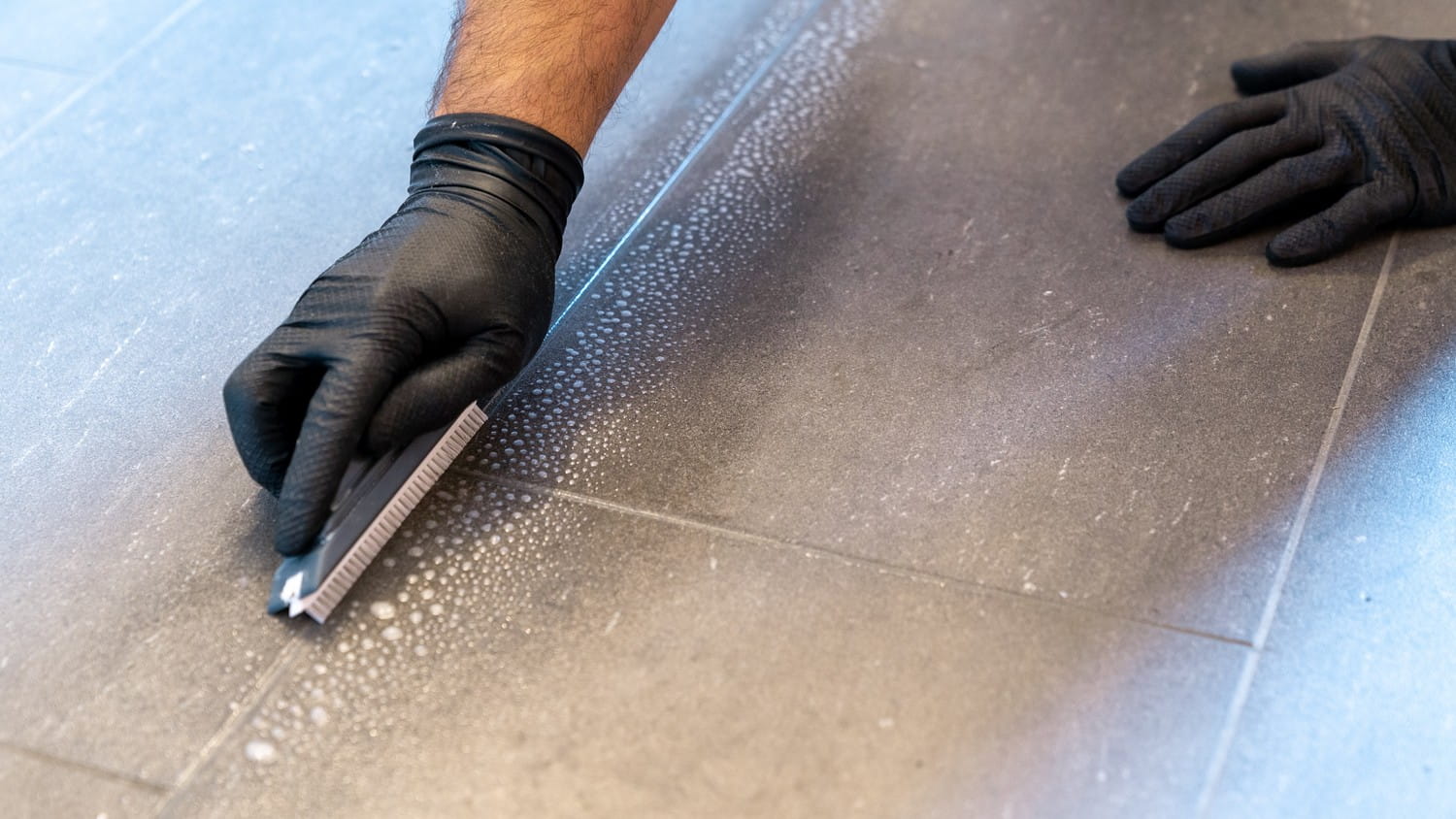 A close-up view of a person's hand holding a scrub brush and cleaning a dirty tile floor.