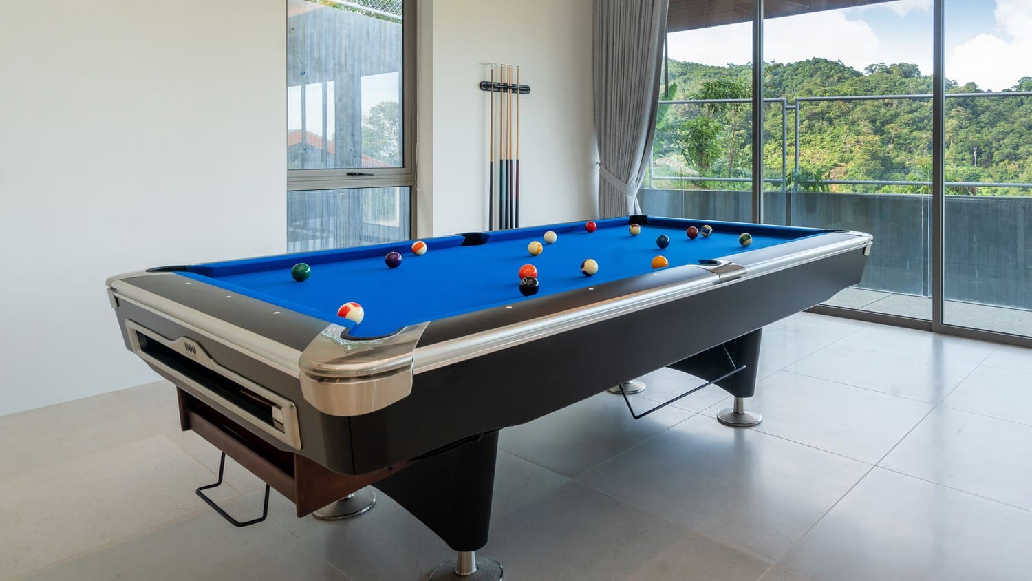 A beautiful pool table with blue felt in a bright room with floor to ceiling glass windows.
