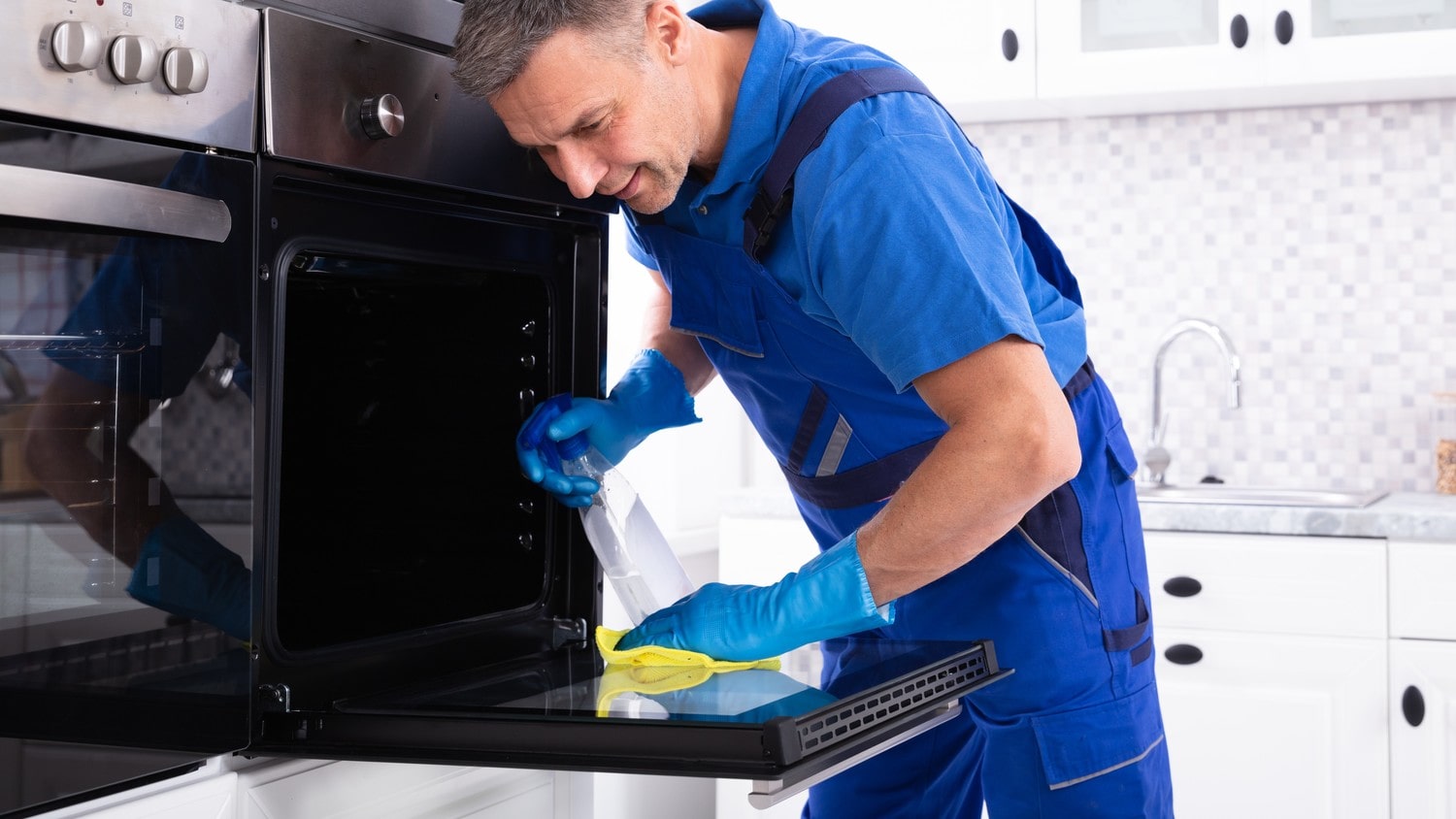 A person wearing rubber gloves and ajumbsuite cleaning the inside of an oven in a brightly lit kitchen.