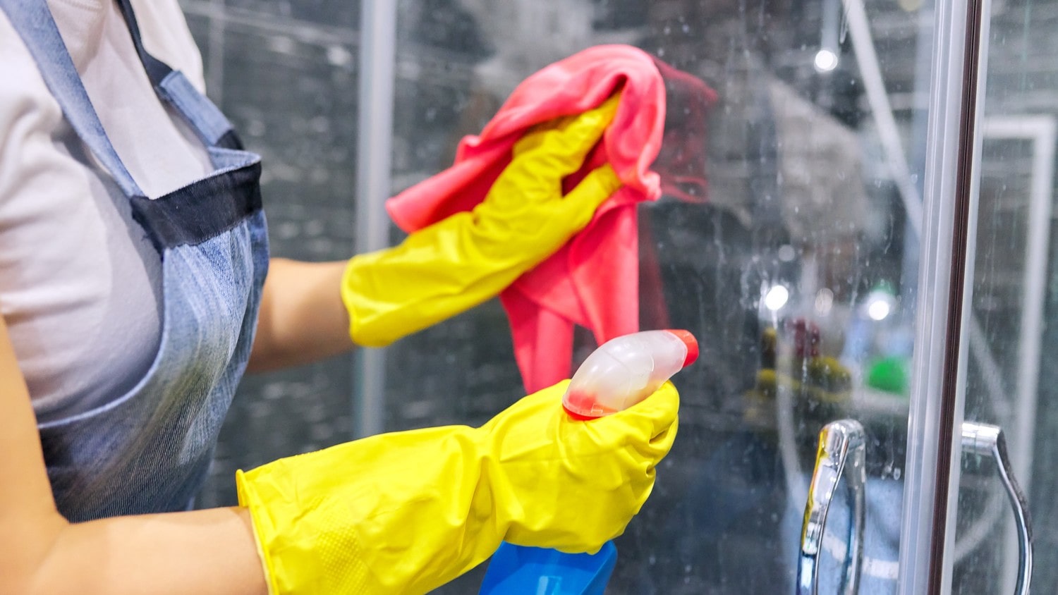 A person cleaning a shower glass with a spray bottle and cleaning clothe.
