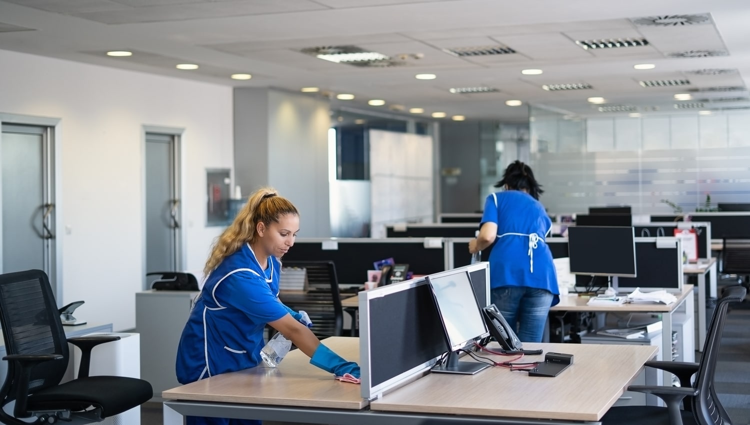 A couple of cleaners in uniform tidying an organized office space with desks, chairs, and computers neatly arranged, ready for a productive workday.