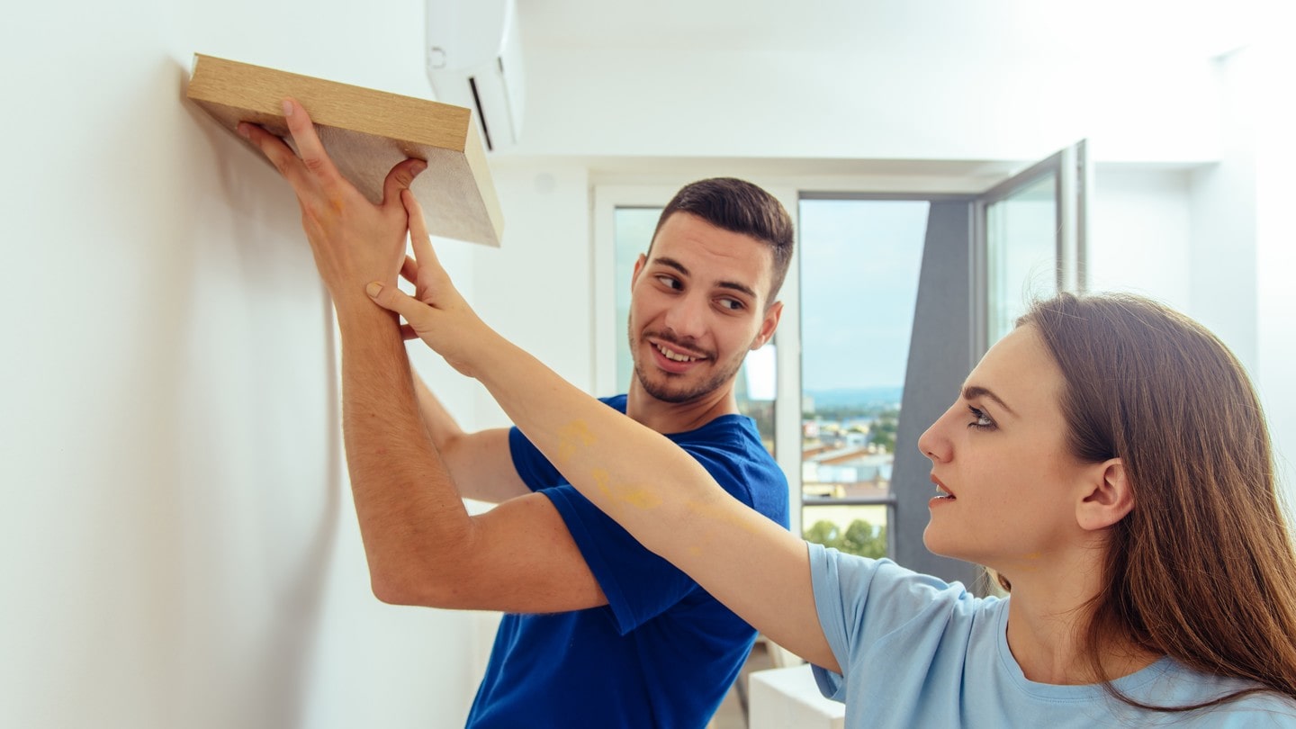 Two people installing a shelf on a white wall.