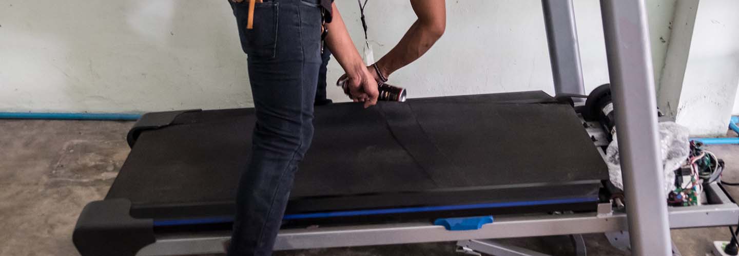 A person repairing a treadmill, focused on fixing the belt and adjusting the tension, with tools and parts scattered around.