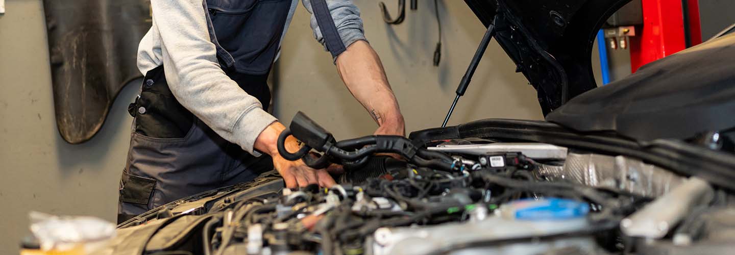 A mobile mechanic working on a car engine, standing under the hood wearing a protective apron over his gray shirt.