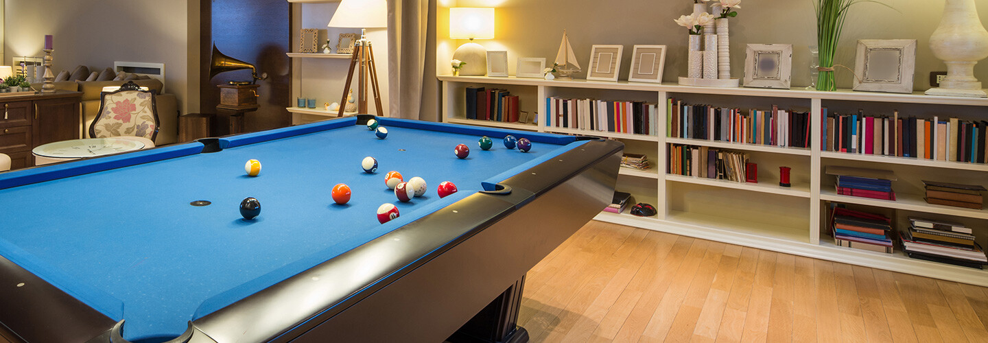 A pool table with blue felt and different billiard balls on top of it, set in a room full of books and pictures.