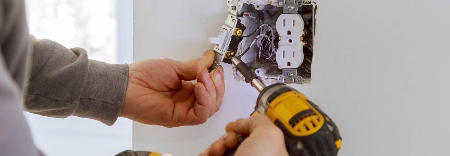 A close-up view of a person's hand installing sockets and switches on a white wall with electrical wires visible.