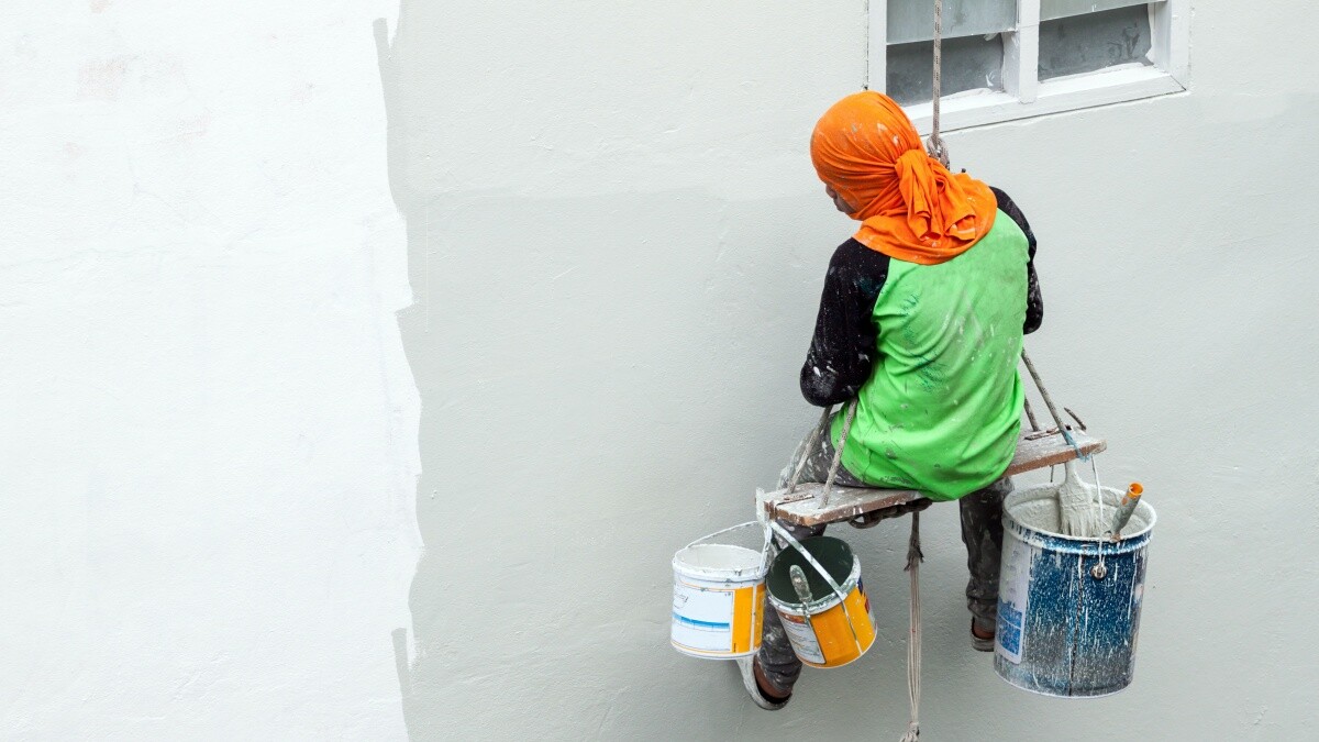 man sitting on makeshift chair hanging from rope painting exterior walls