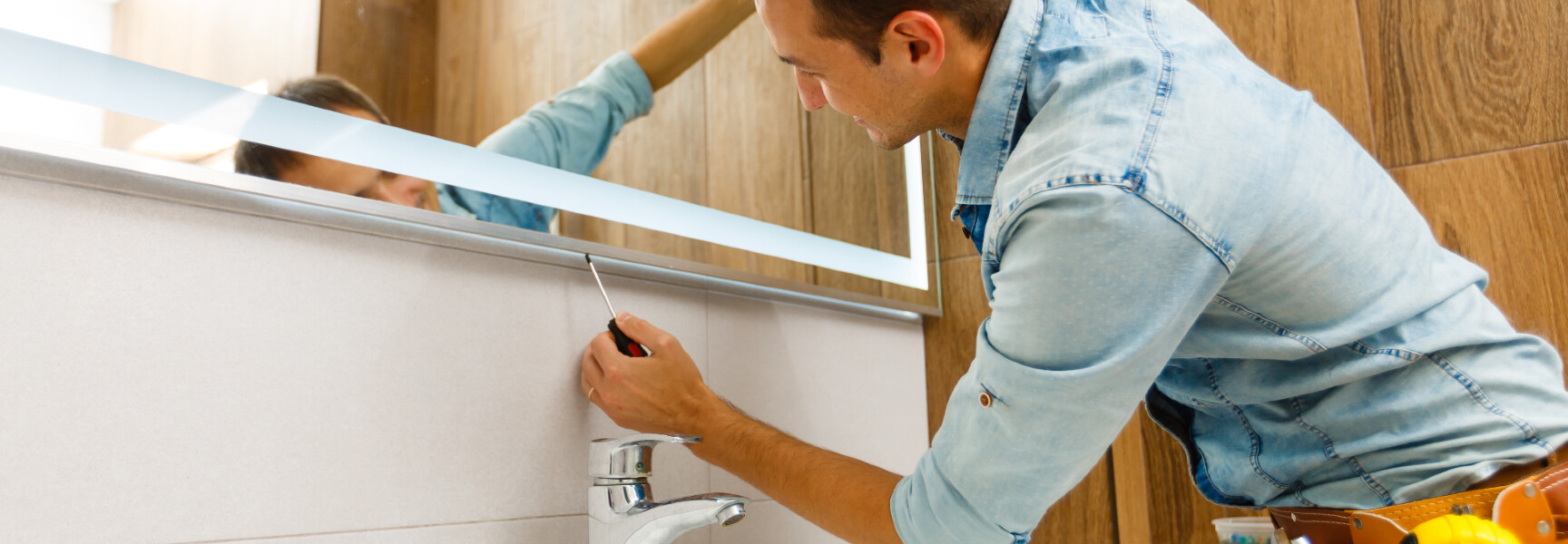 a handyman in a blue denim shirt making repairs on a mirror above the sink.