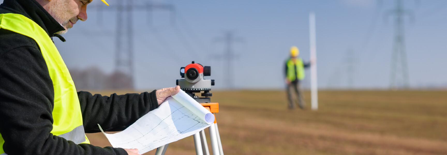 A person in a reflective vest and hard hat holding a surveying tool, measuring the land with a beautiful sunset in the background.