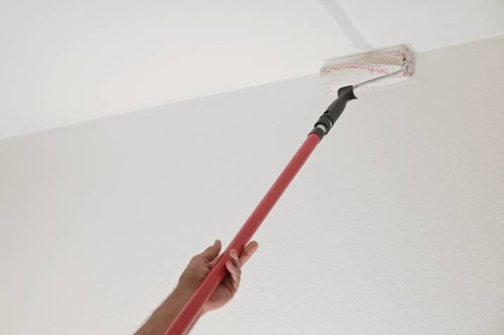 a hand holding a paint roller on a long telescopic pole to paint the ceiling in a house