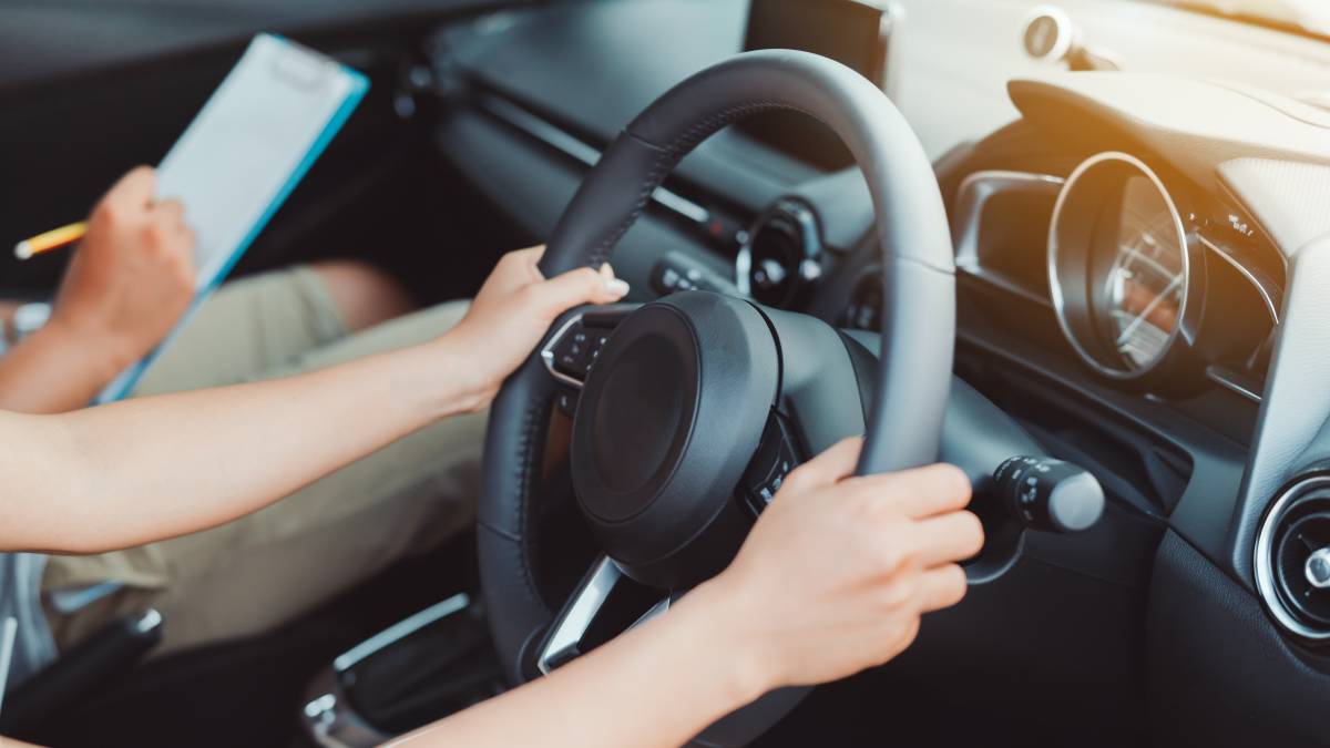 A person gripping a steering wheel during a driving lesson, with an instructor holding a clipboard.