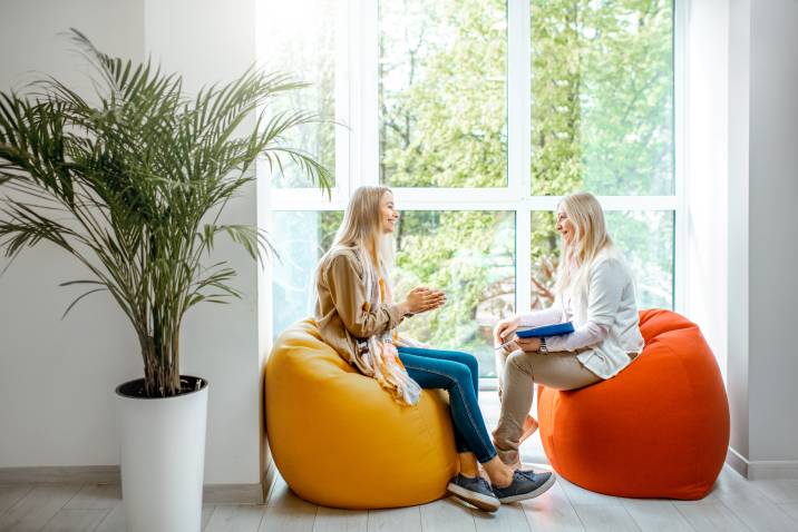 Clinical social work. Senior female psychologist with young woman. Sitting on comfortable chairs beside floor-to-ceiling window during counselling