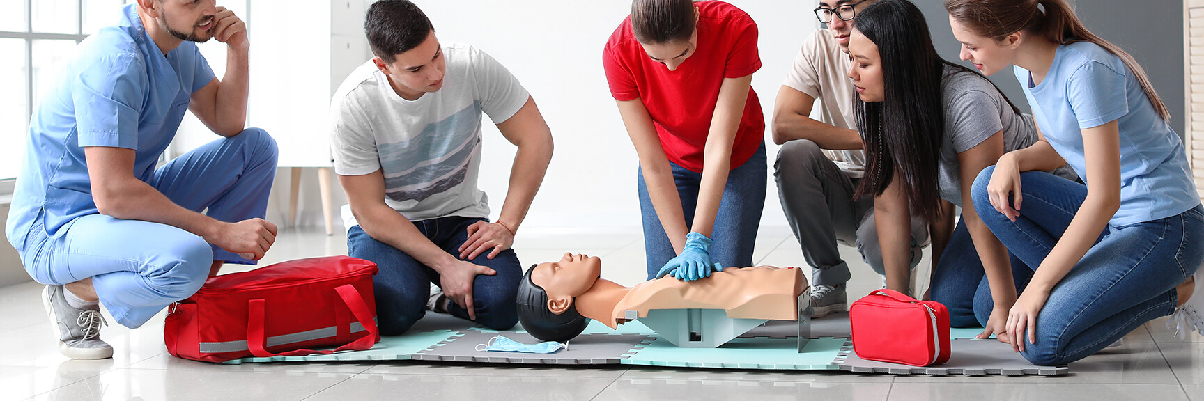 A group of people participating in CPR training, with one person performing chest compressions on a mannequin while others observe and learn.