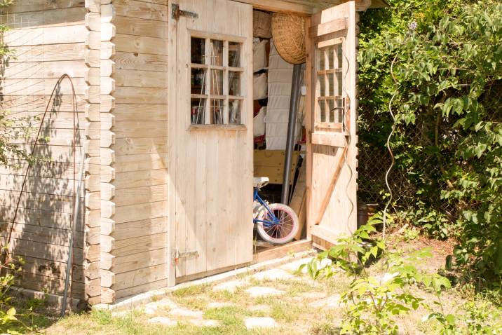 A wooden log cabin shed with open doors, revealing stored items like a bicycle, surrounded by greenery and a sunny outdoor setting