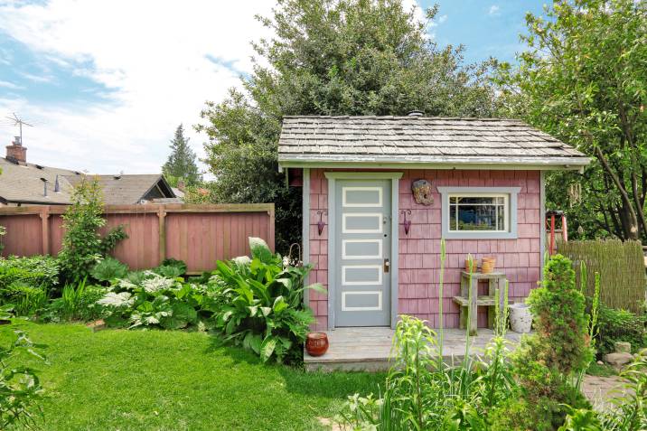 A small backyard barn painted in pink with a white door, surrounded by vibrant plants and trees