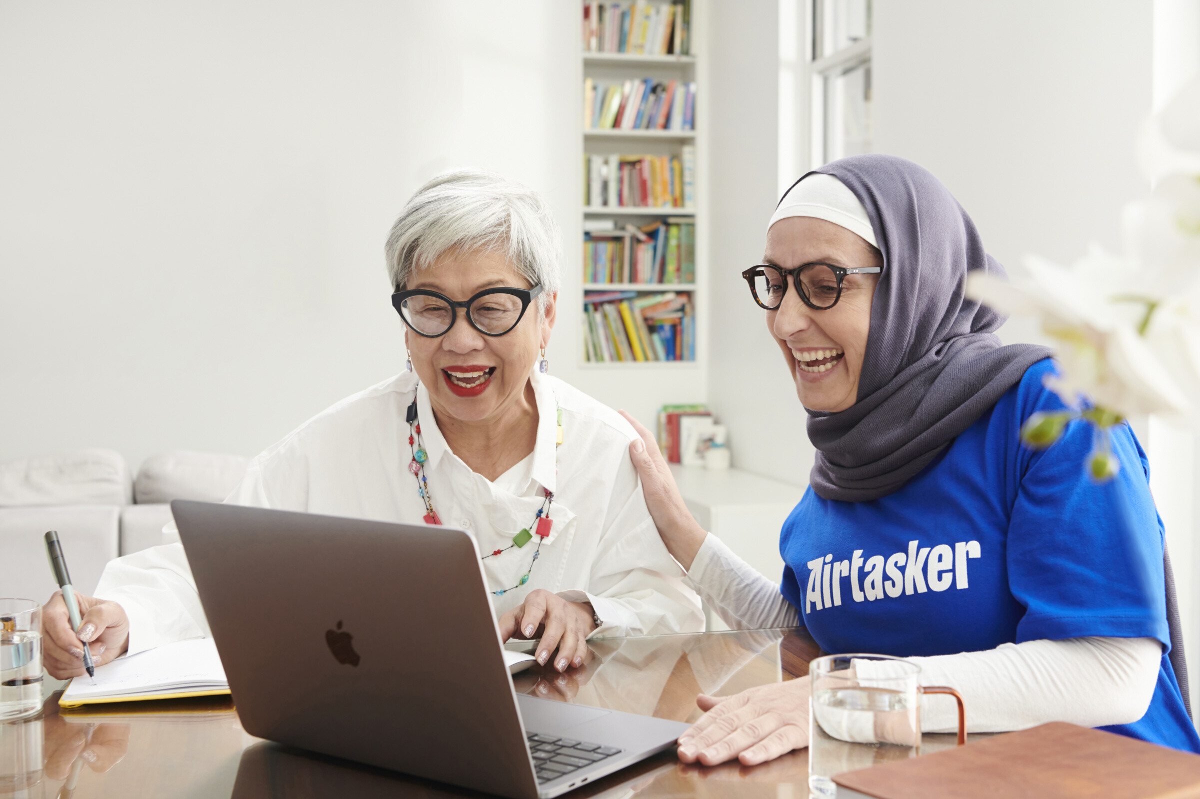 An elderly woman sitting at a desk, being taught how to use a laptop.