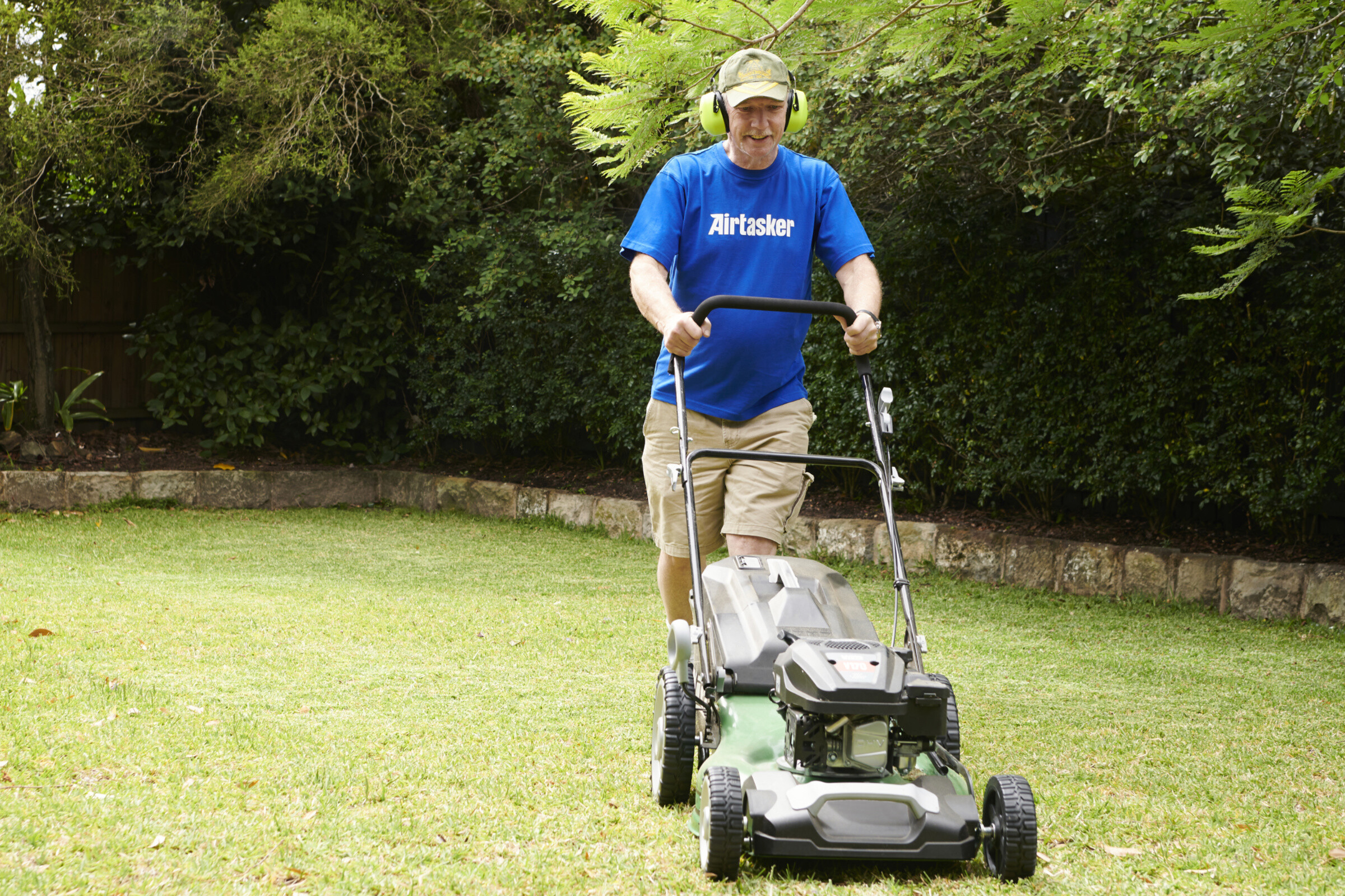 A neatly mowed lawn with vibrant green grass, surrounded by colorful flowers and a person using a lawnmower in the background.