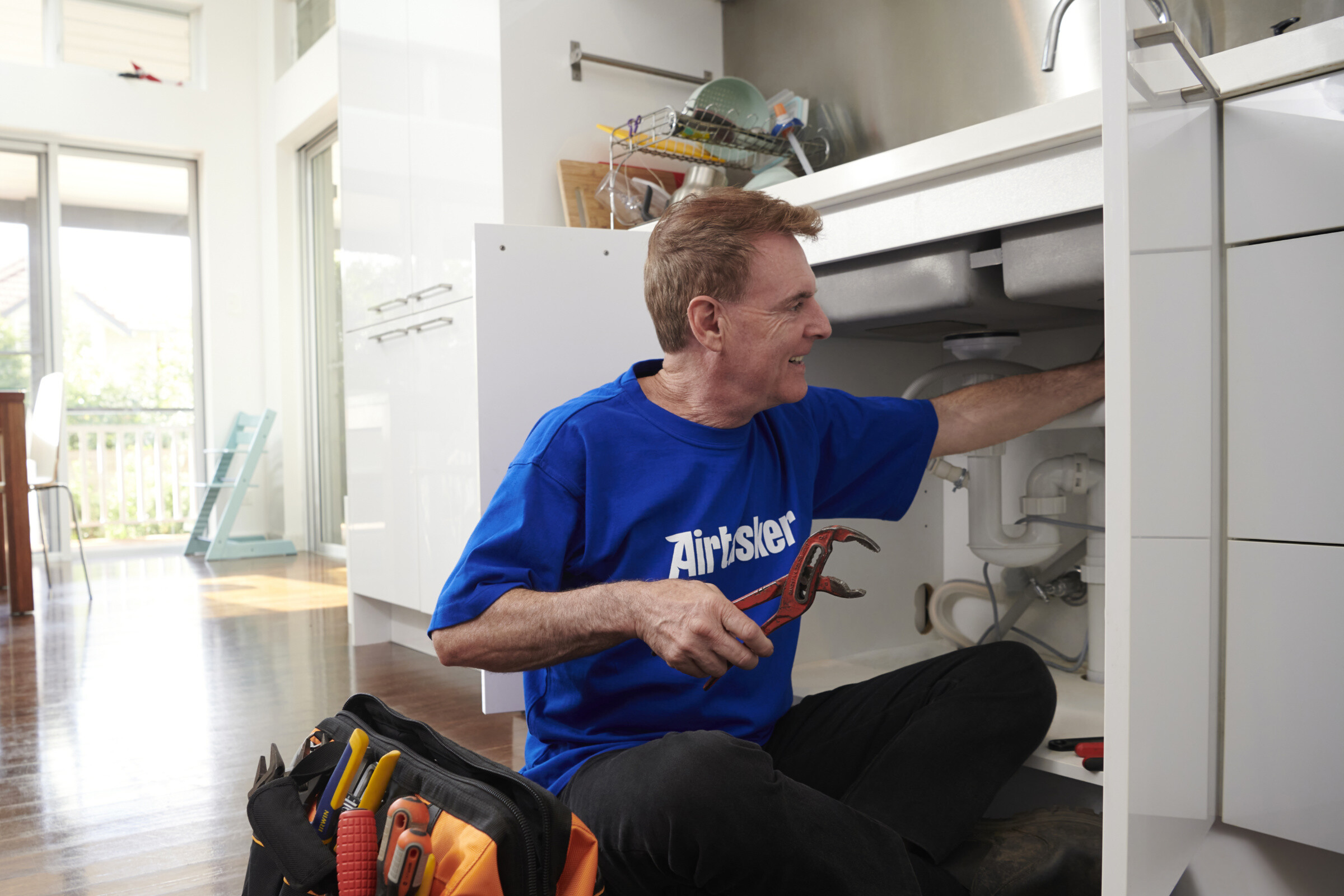 A plumber working on a sink, holding a wrench, with tools and pipes in the background.