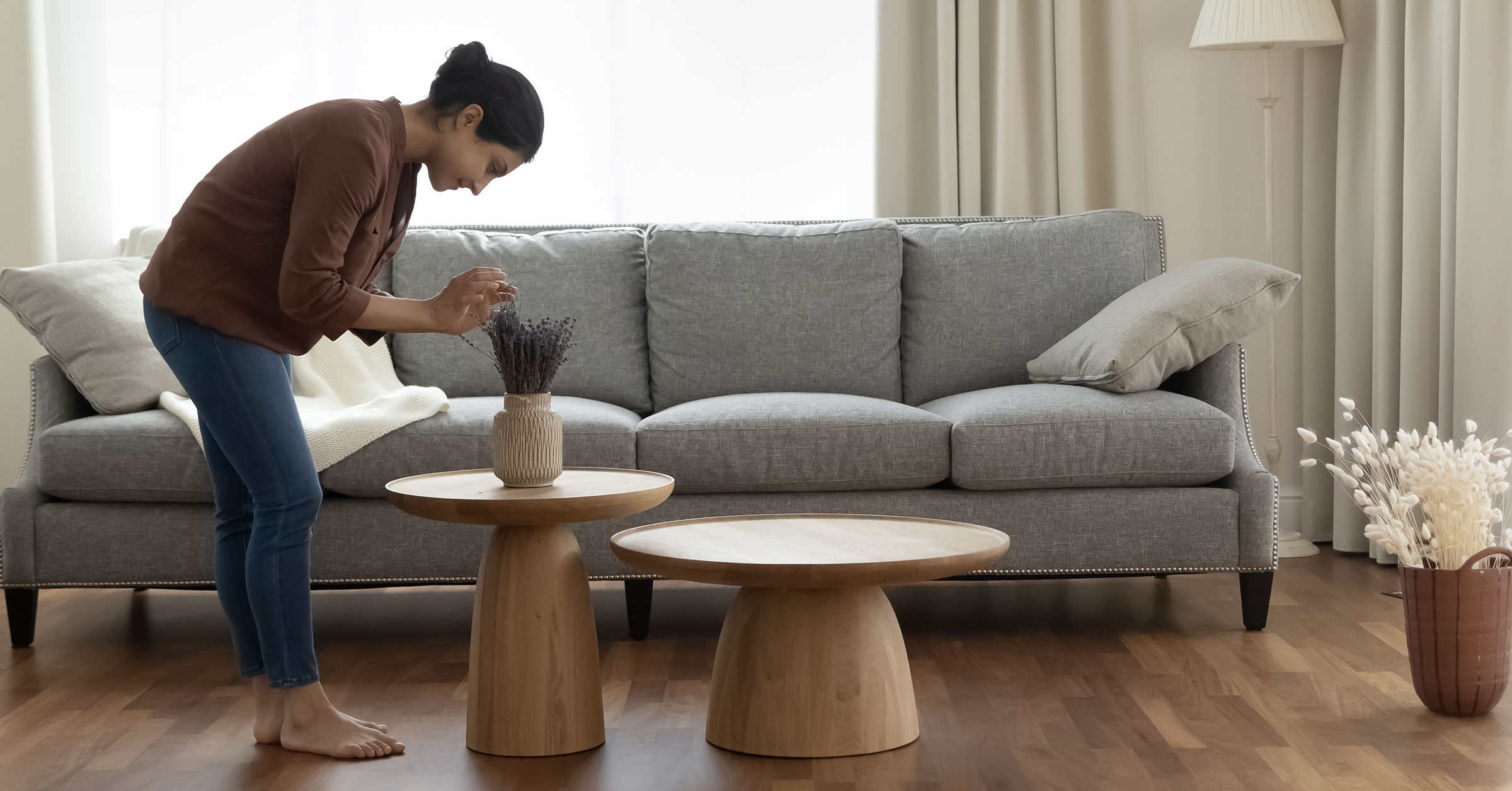 An interior designer placing flowers in a vase on top of a brown, minimalist table in a well lit living room.