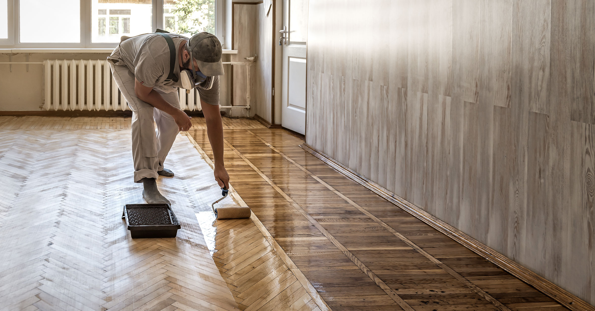A flooring professional varnishing the wooden floors of a spacious and well lit room.