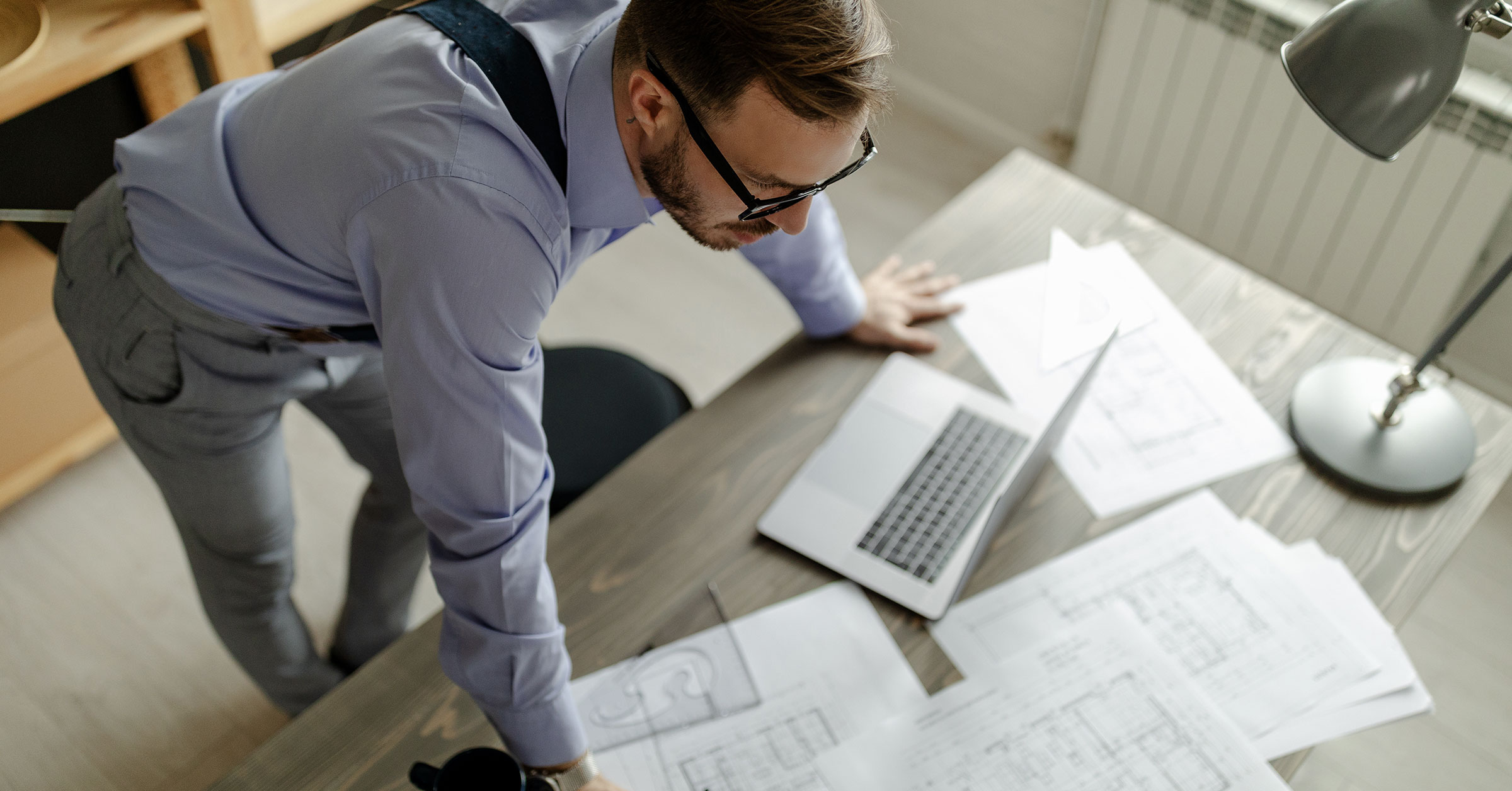 A top view of a draftsman in a suit, woking with blueprints on his office desk.