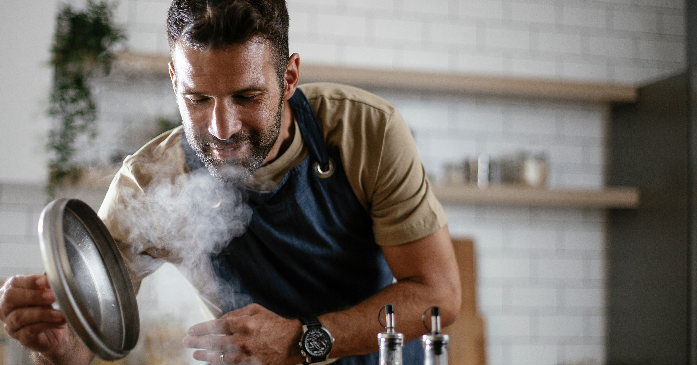 A close up photo of a chef holding a pot cover, checking on a dish as it cooks on a stove.