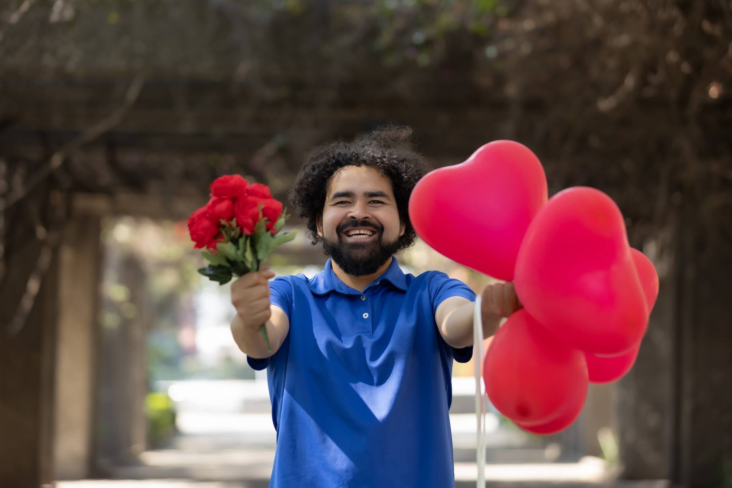 A delivery guy wearing a blue shirt and carrying personalised heart-shaped balloons on one hand and red flowers on the other