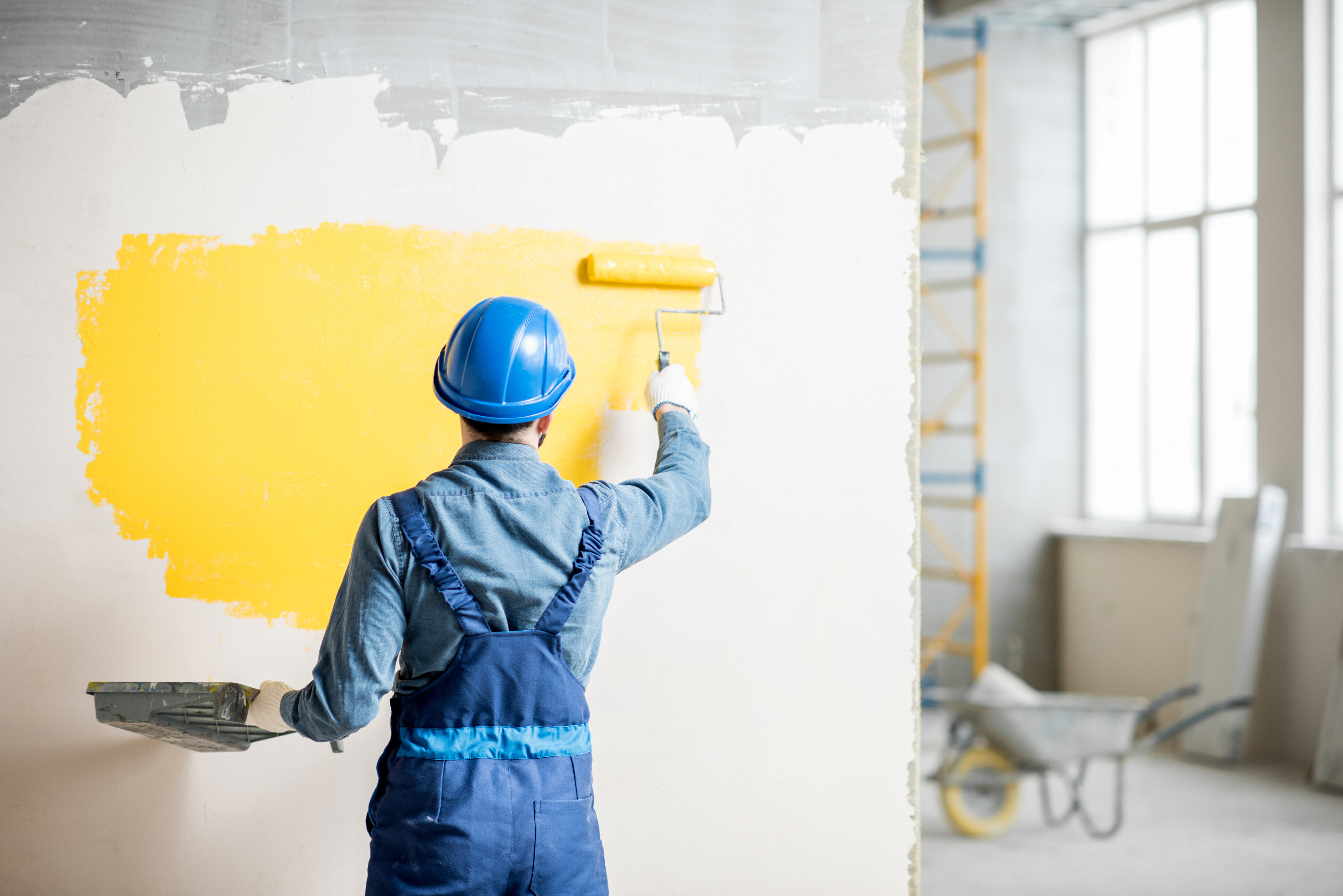 A professional painter wearing a blue jumpsuit painting the wall of an office space.