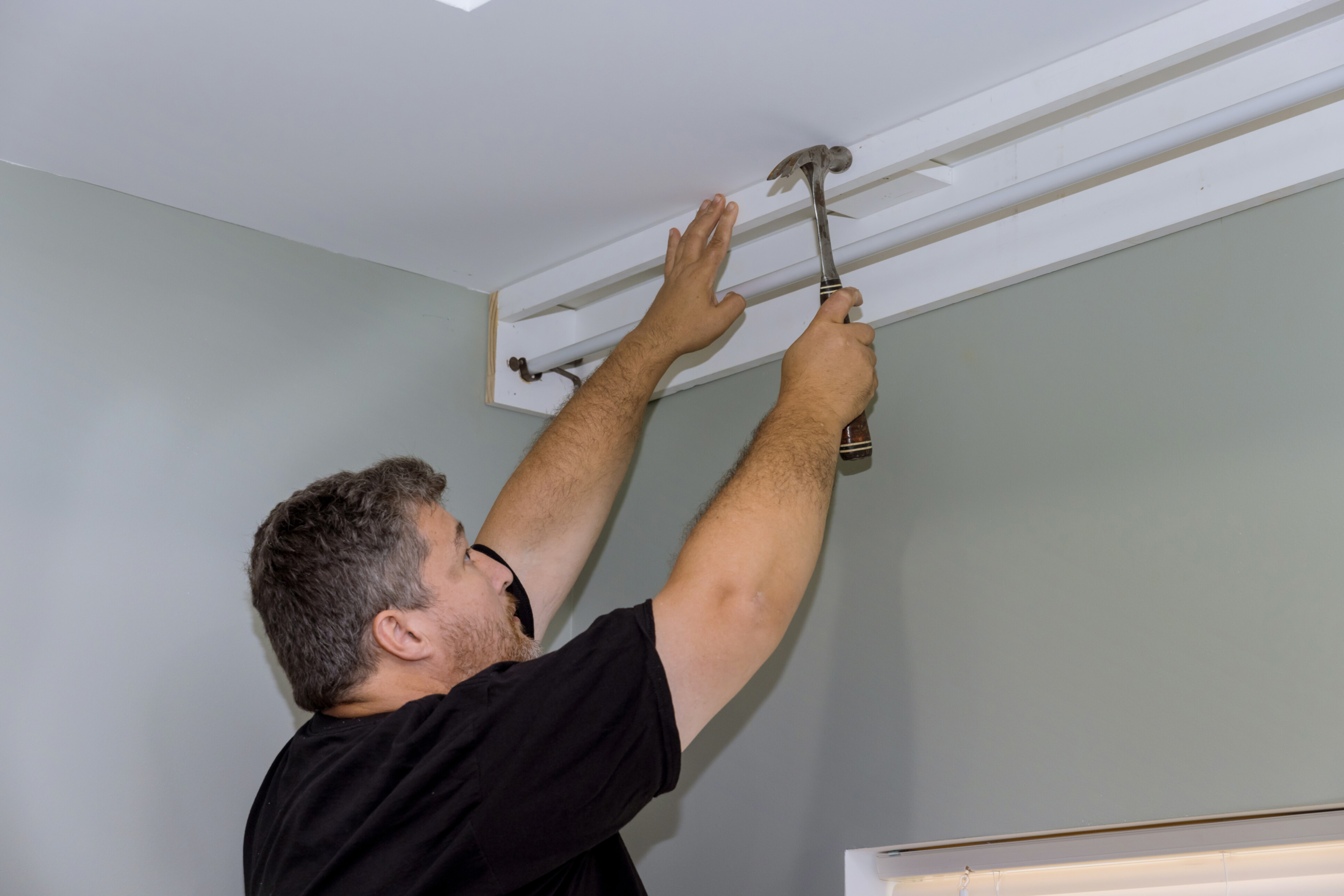 A handyman installing a curtain rod and wood cornices over a window.