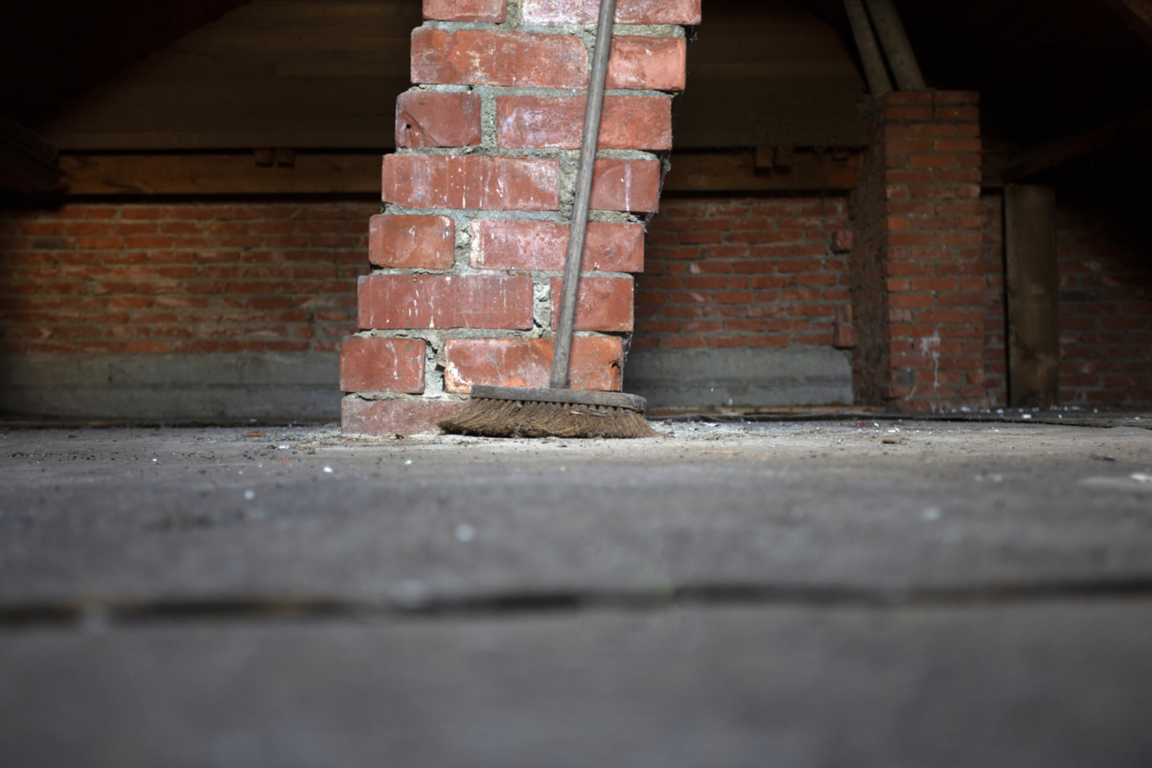 A used broom leaning against a brick post in a dirty attic.