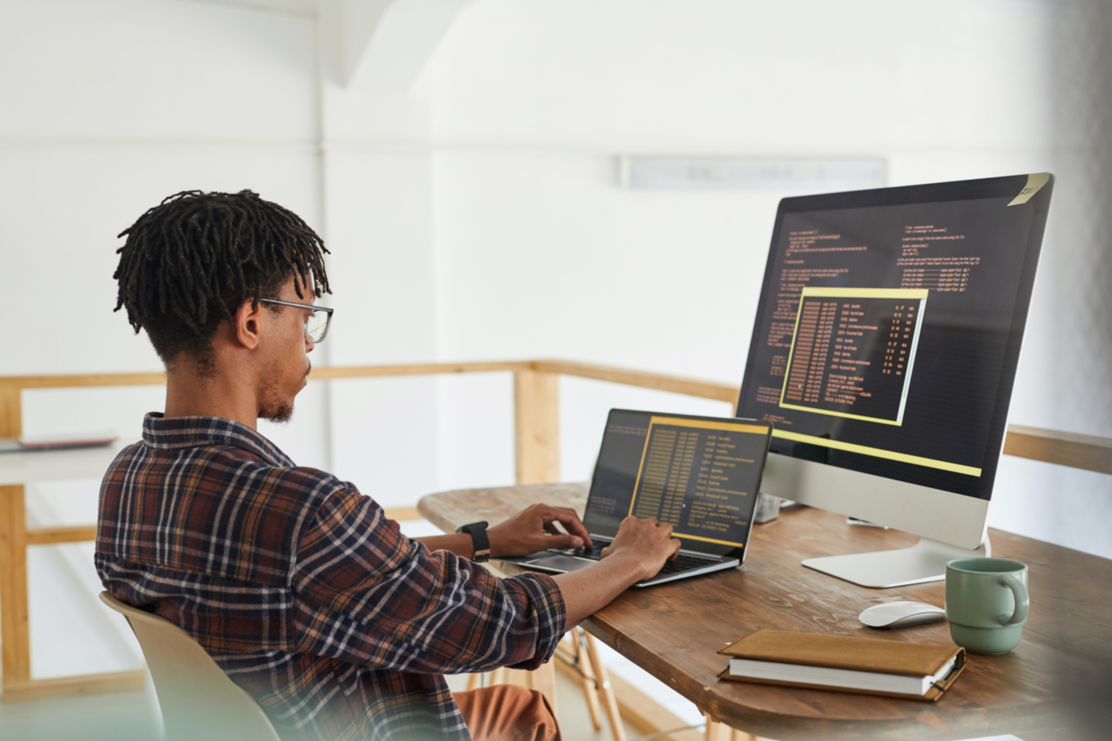 An african american man coding in front of 2 screens on his desk.