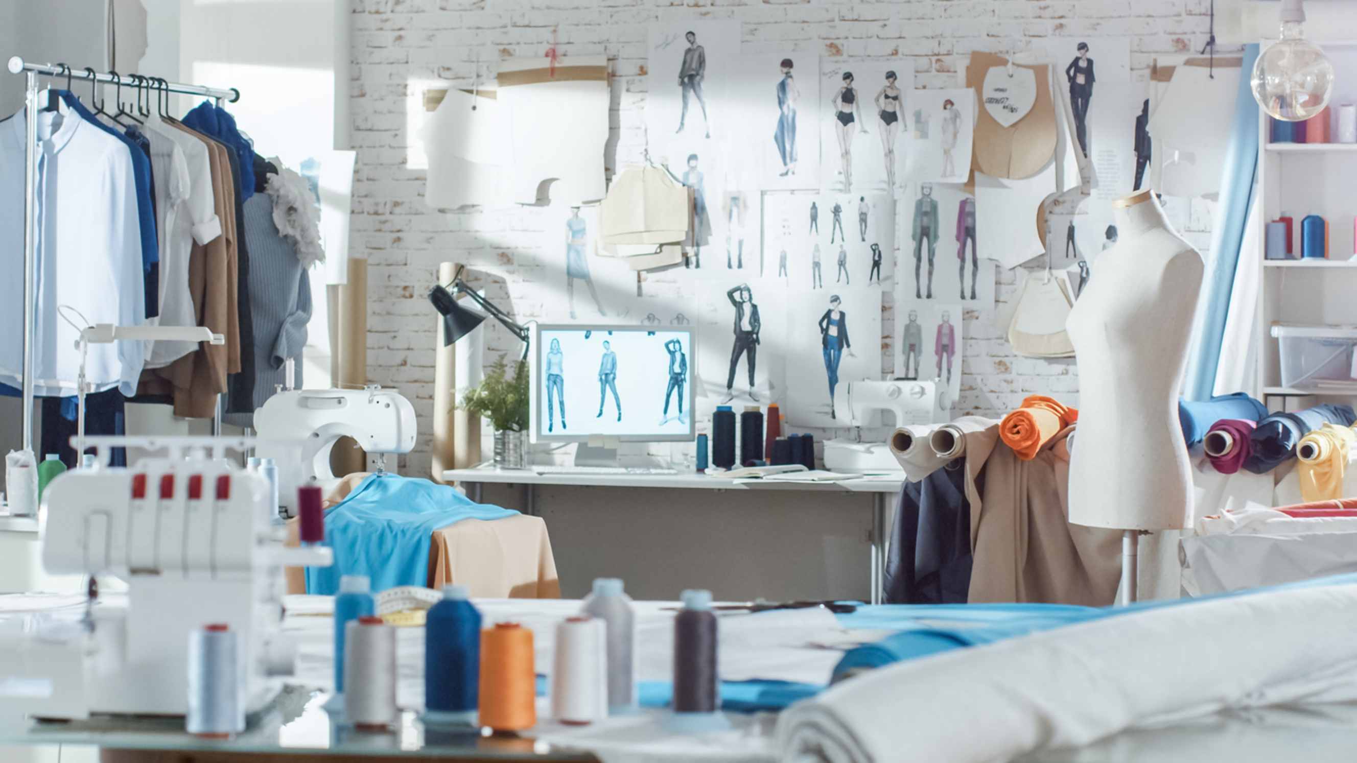 Hand sewing vs machine - Shot of a fashion design studio with hanging clothes, a sewing machine, various sewing essentials on the table, mannequins standing, and fabrics.