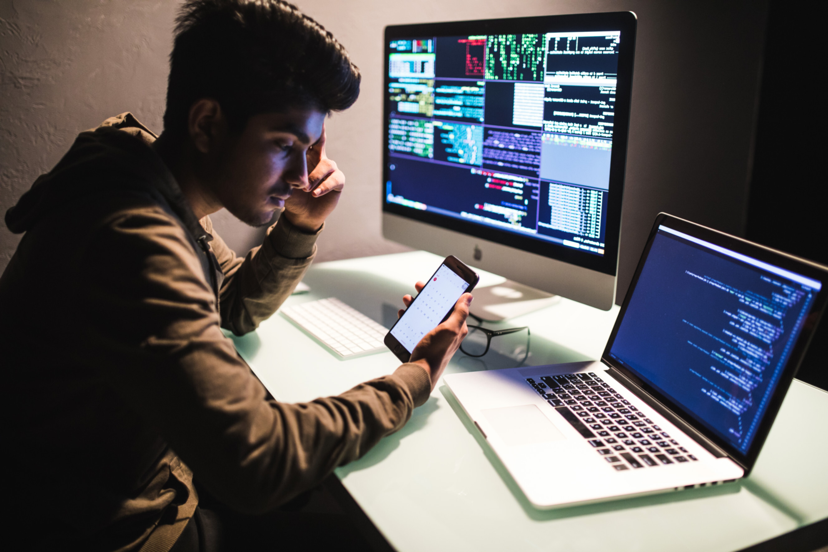 App developer near me reviewing mobile app prototype on smartphone, with laptop and external monitor displaying source code and terminal commands.