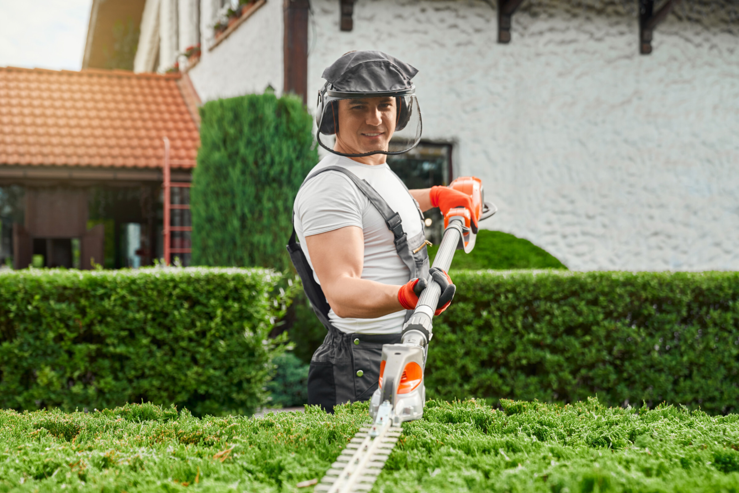 Professional gardener using an electric hedge trimmer to neatly trim hedges in front of a residential home 