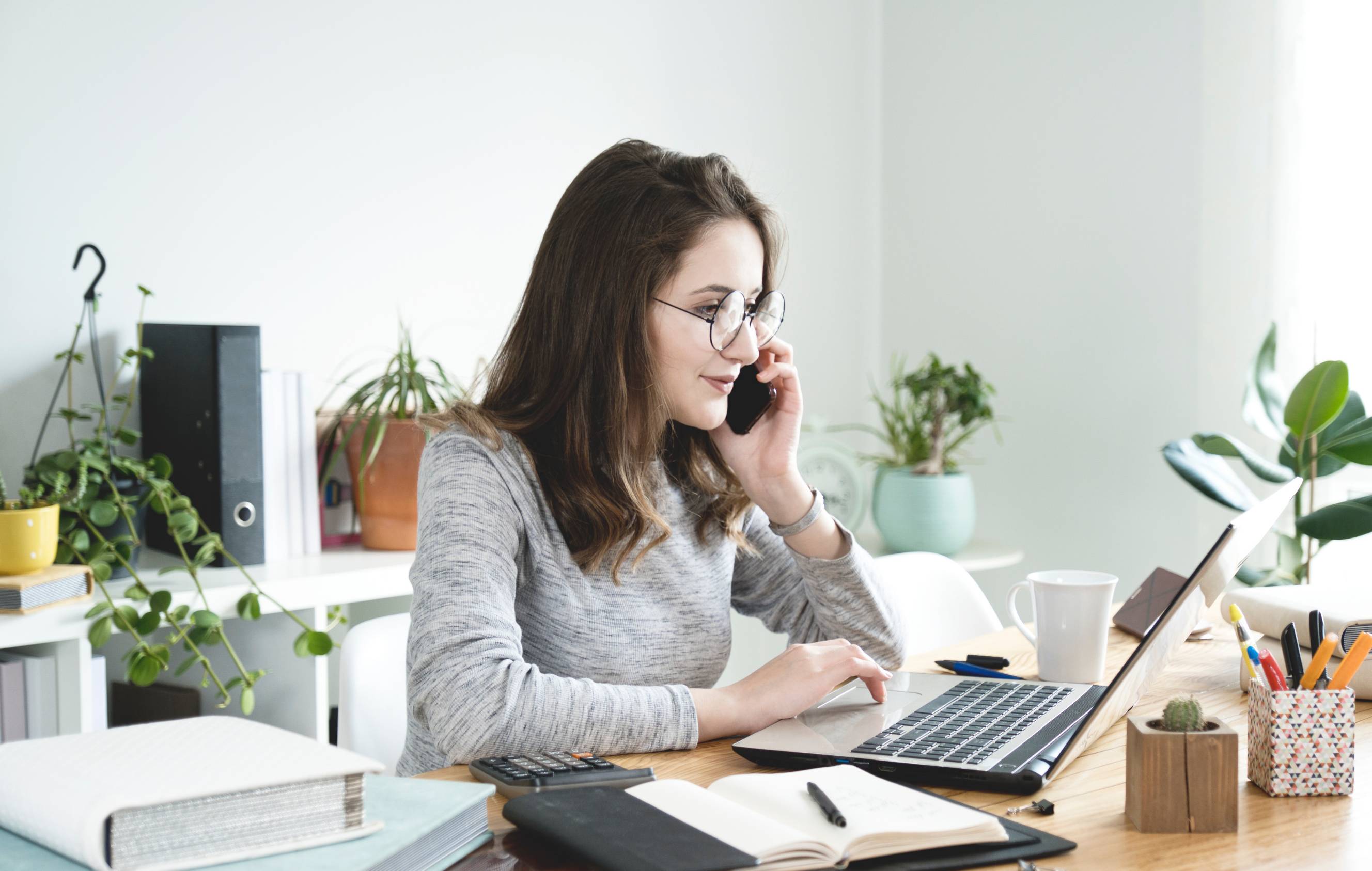 An office and admin worker talking on the phone while using a laptop at a desk for clerical tasks.