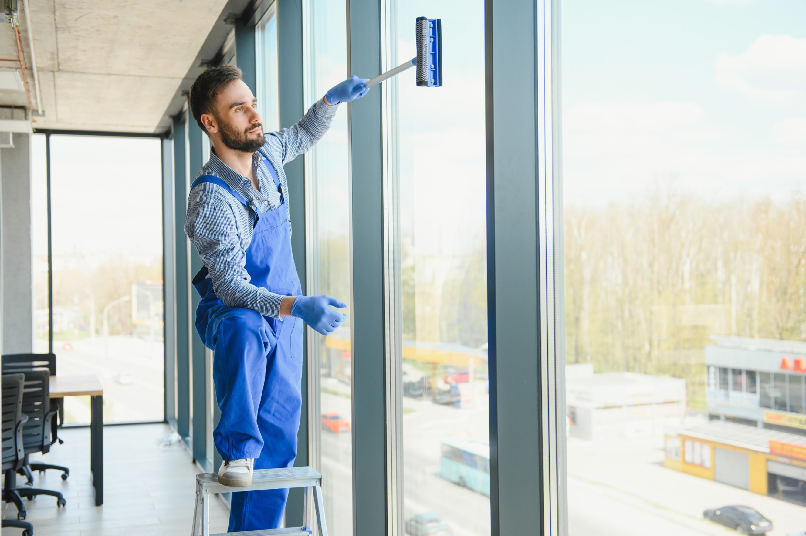 Professional cleaner in blue overalls using a squeegee to clean large office windows, representing commercial window cleaning near me services.