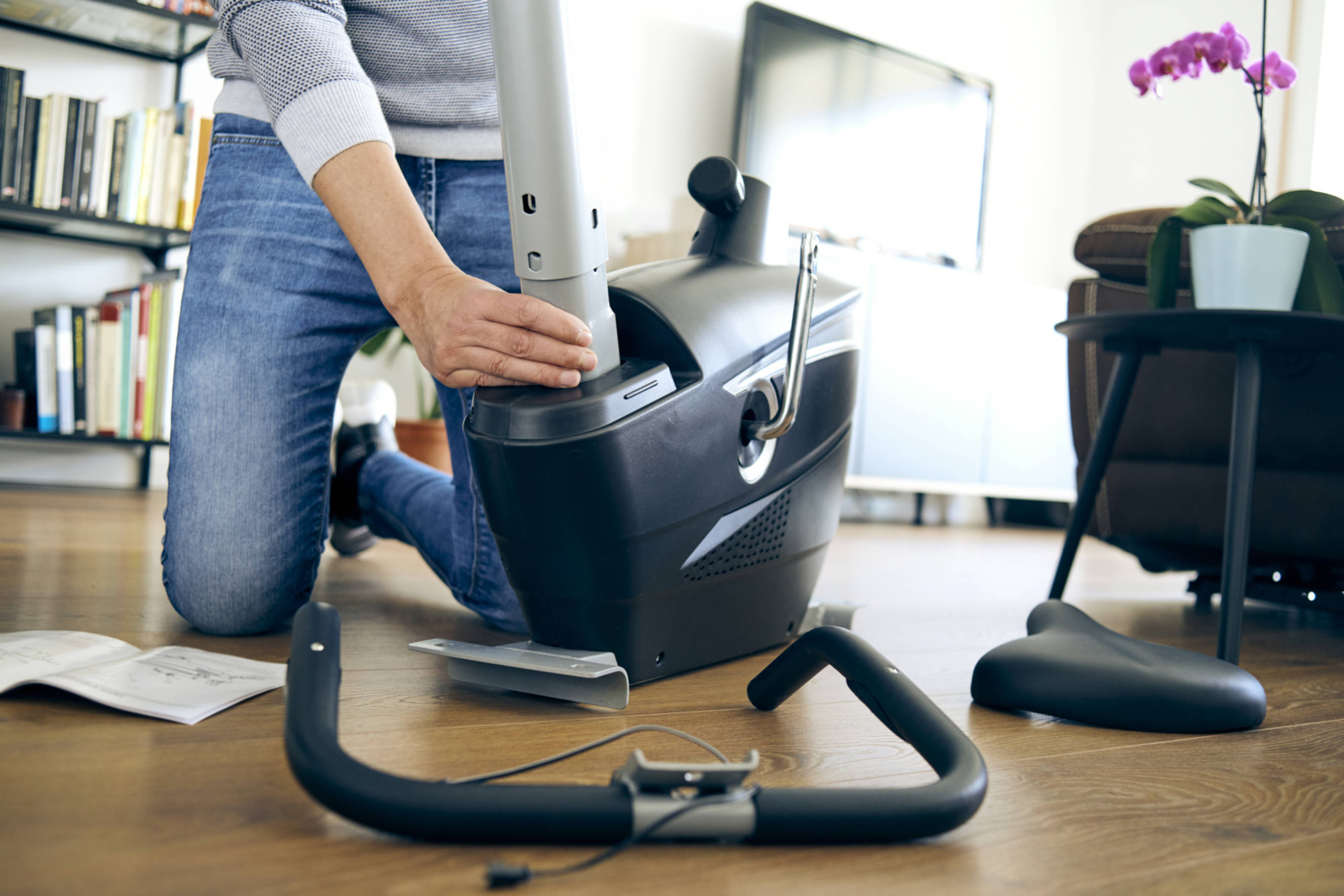 Person assembling an exercise bike in a modern living room, highlighting the step-by-step process of exercise bike assembly. The scene includes a close-up of the bike’s base, tools, and instructional manual, showcasing a comfortable home environment.