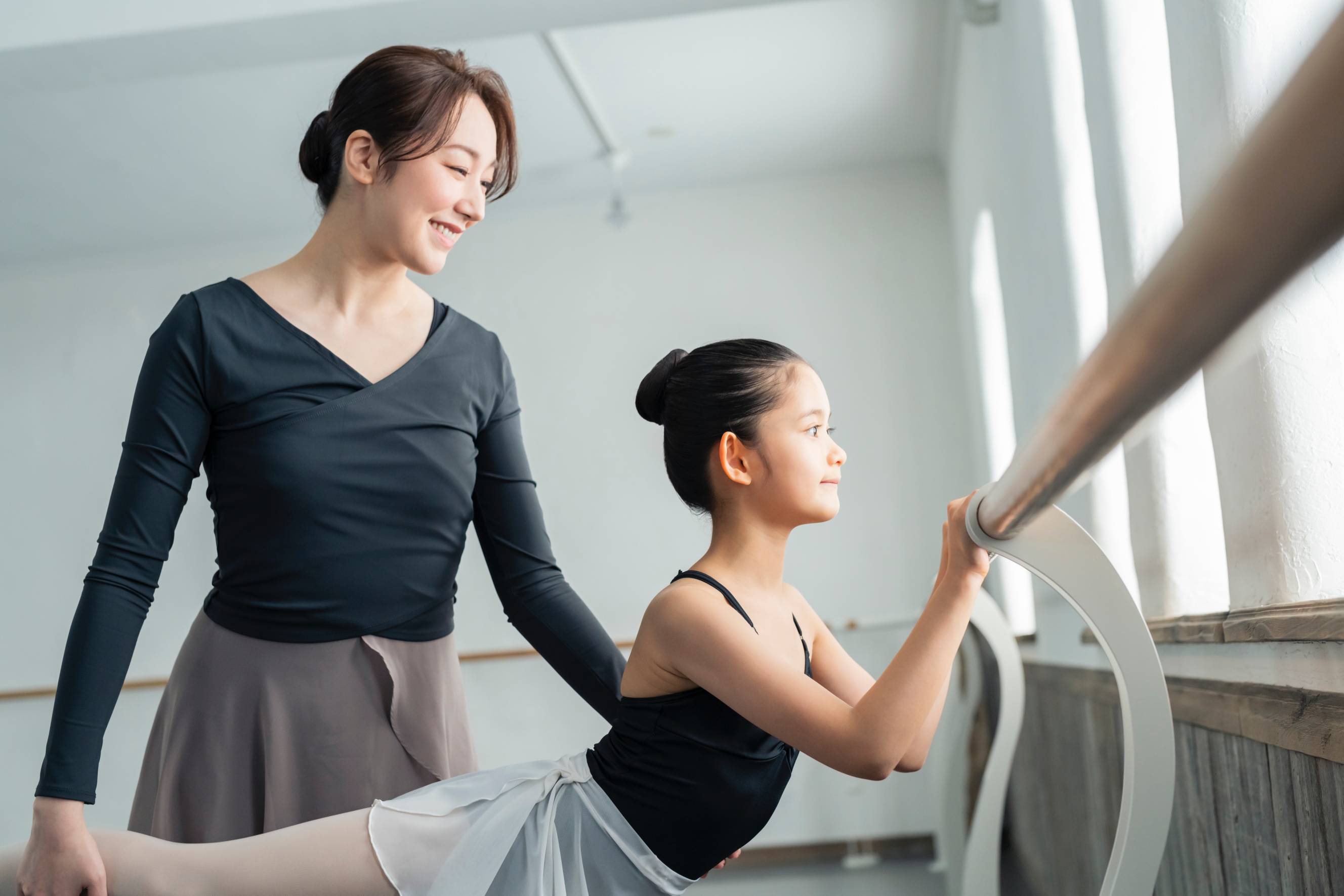 A ballet teacher with a young female student practising ballet together in a bright studio.