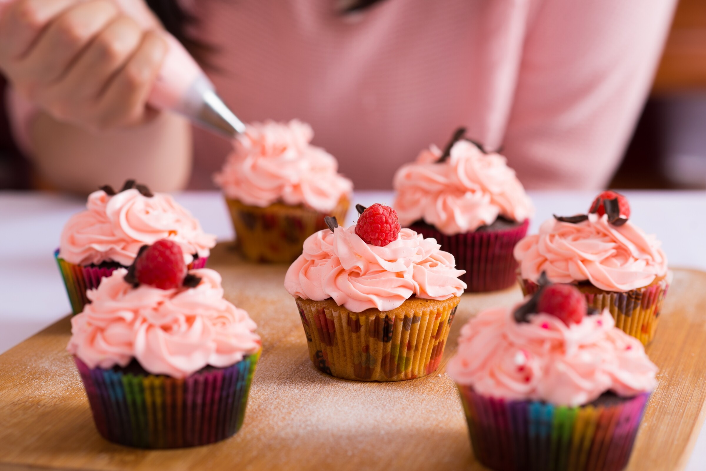 A baker decorating personalised cupcakes with pink frosting and raspberries, featuring colorful cupcake liners on a wooden board.