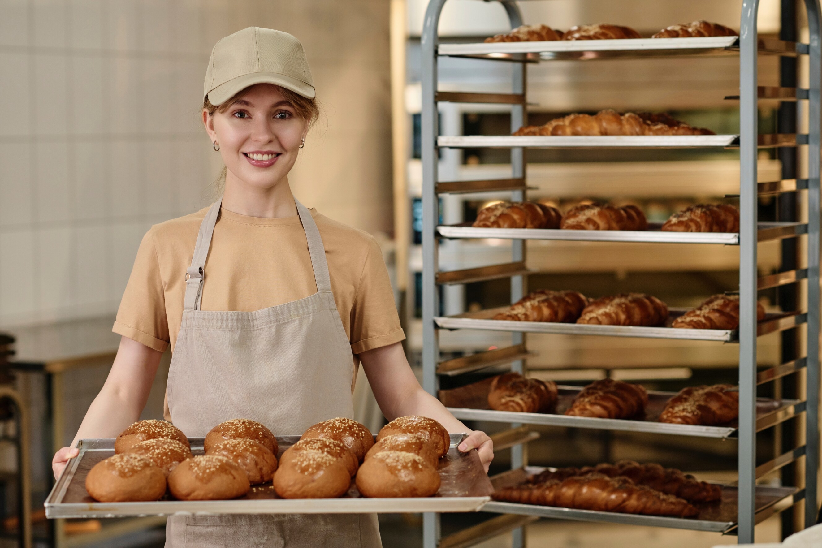 Smiling artisan baker holding a tray of freshly baked bread rolls in a bakery, with racks of bread in the background.