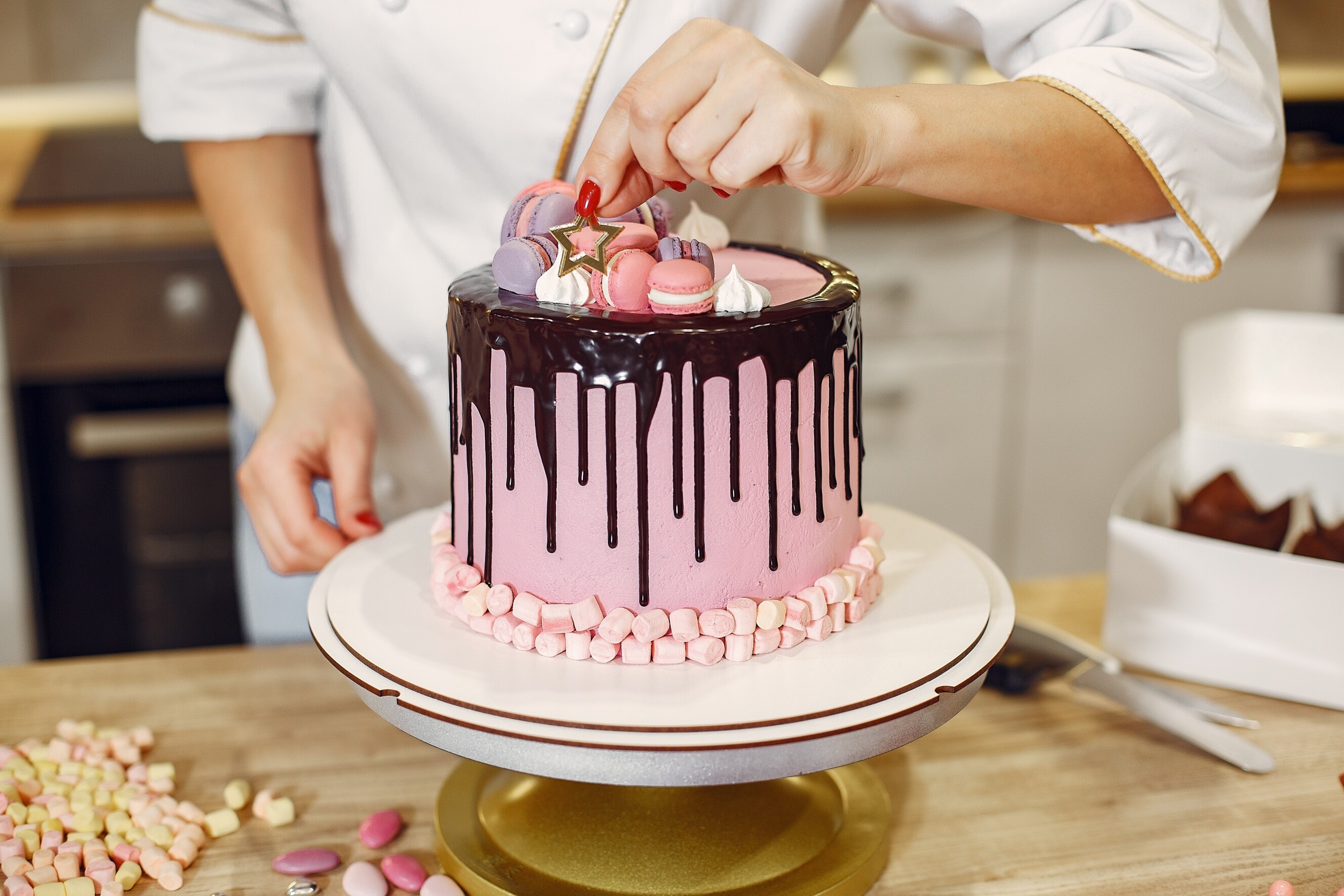 Custom cake baker decorating a pink cake with macarons, marshmallows, and chocolate drips in a kitchen.