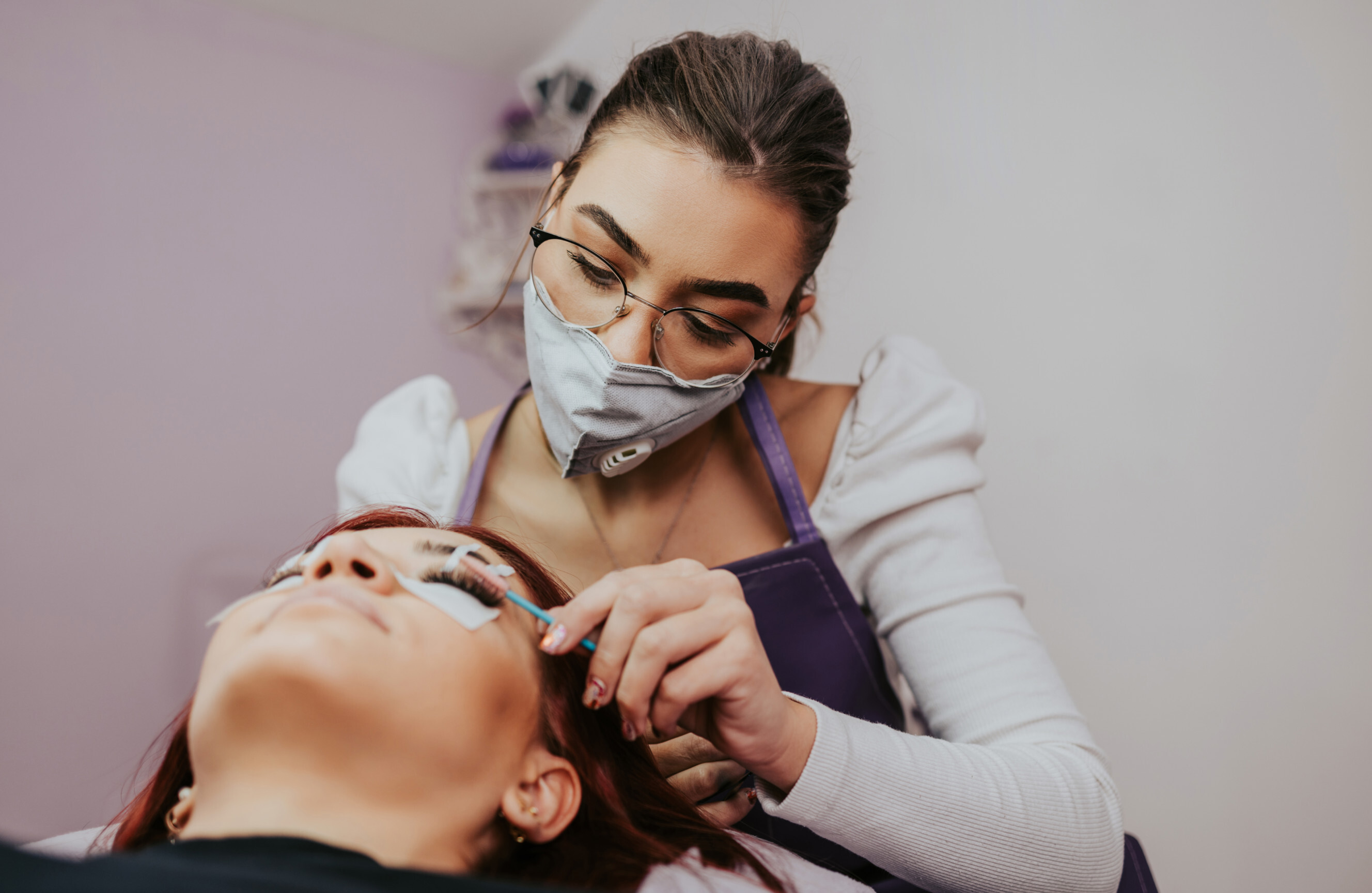 A beauty technician wearing a face mask carefully applies an eyelash perm to a client lying down.