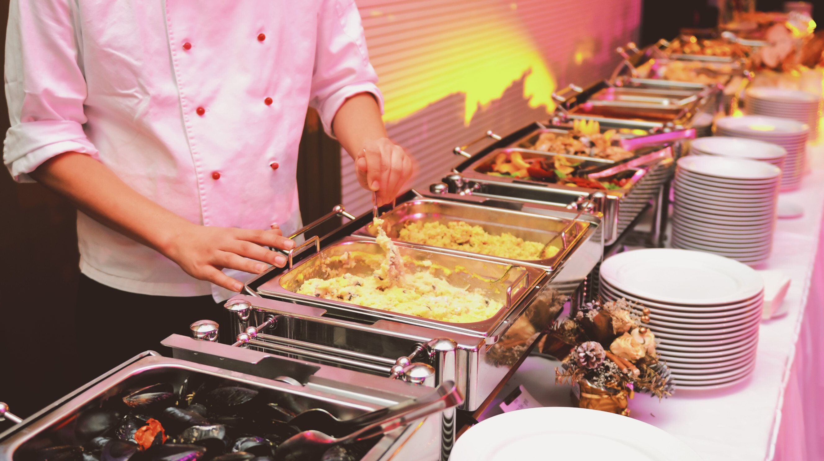 A wait staff preparing food for service behind a buffet table with multiple trays of food and plates on top.