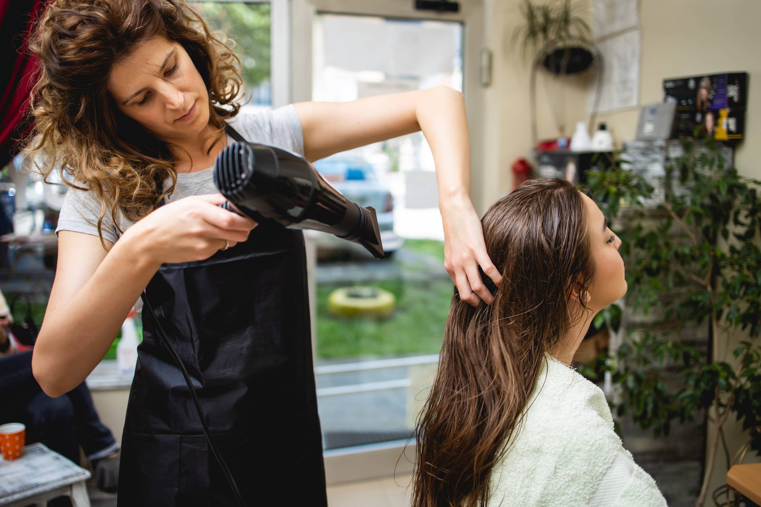 A female hair dresser blow drying a female client's long brown hair at a salon. 