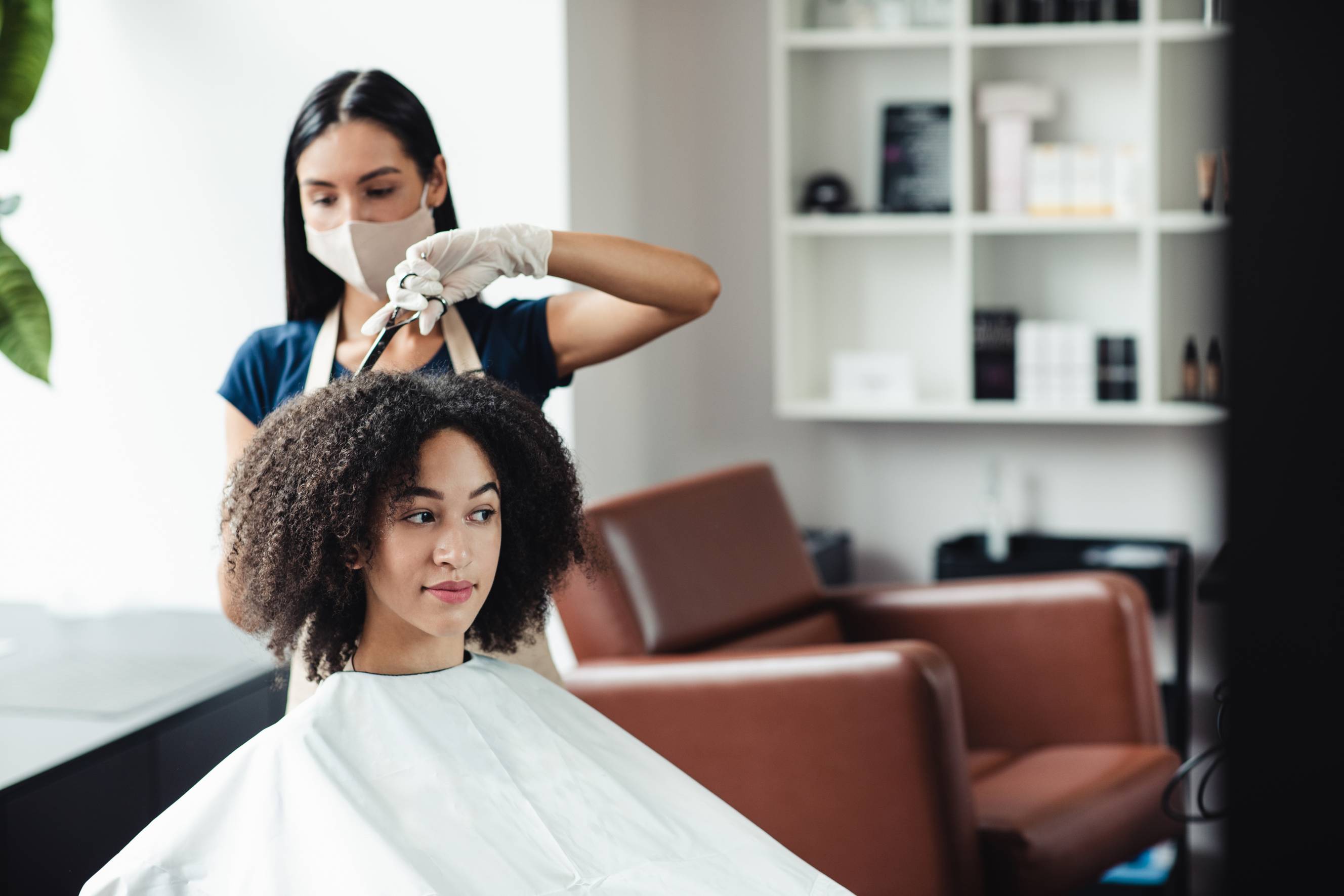 A female curly hair specialist with long dark hair, wearing a mask, styling the hair of another woman with curly, afro-textured hair. 