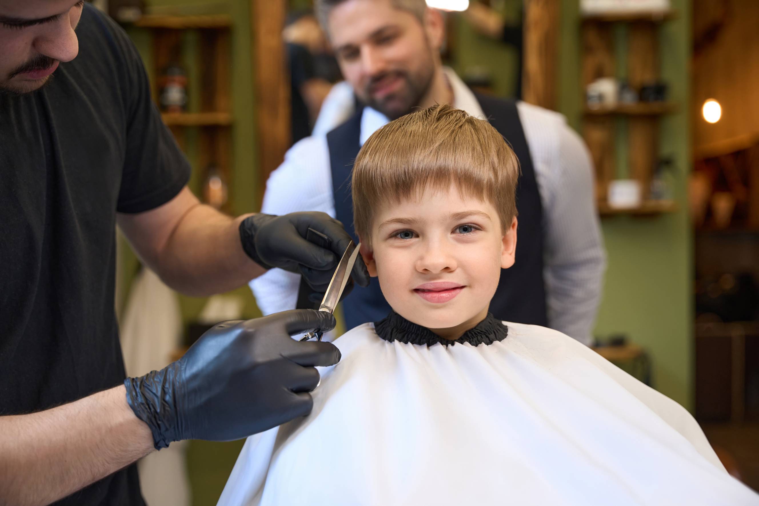 A hairstylist cutting a young boy's hair in a traditional barbershop setting, with a smiling client in a white cape in the background.