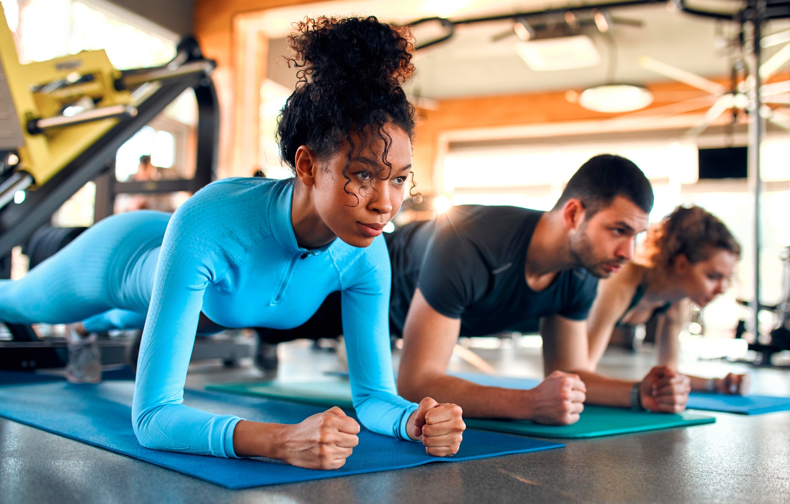 Fit woman in blue sportswear doing a plank exercise on a yoga mat, while other people exercise in a gym setting behind her.